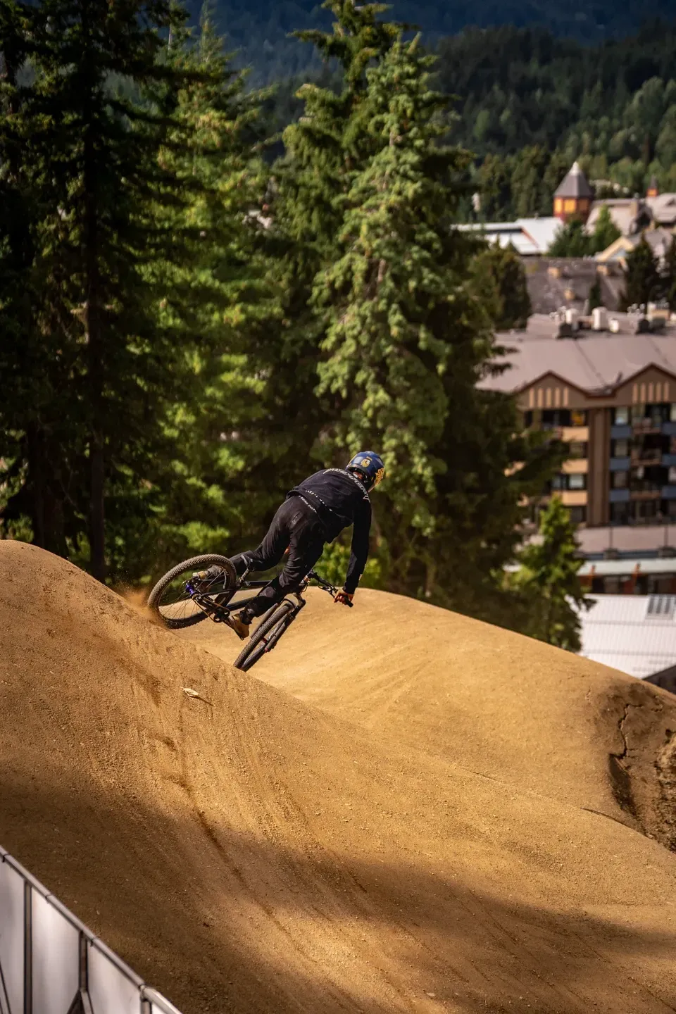 Mountainbiker legt sich auf einer Lehmstrecke in die Kurve, im Hintergrund Wald und Gebäude eines Bergorts