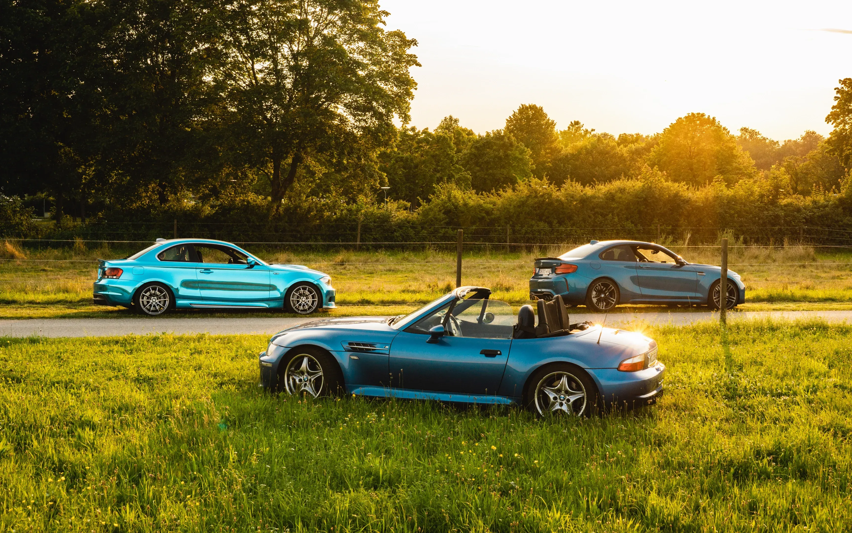 Drei blaue Sportwagen, vorne ein offener Roadster, stehen im goldenen Abendlicht auf einer Wiese am Feldweg