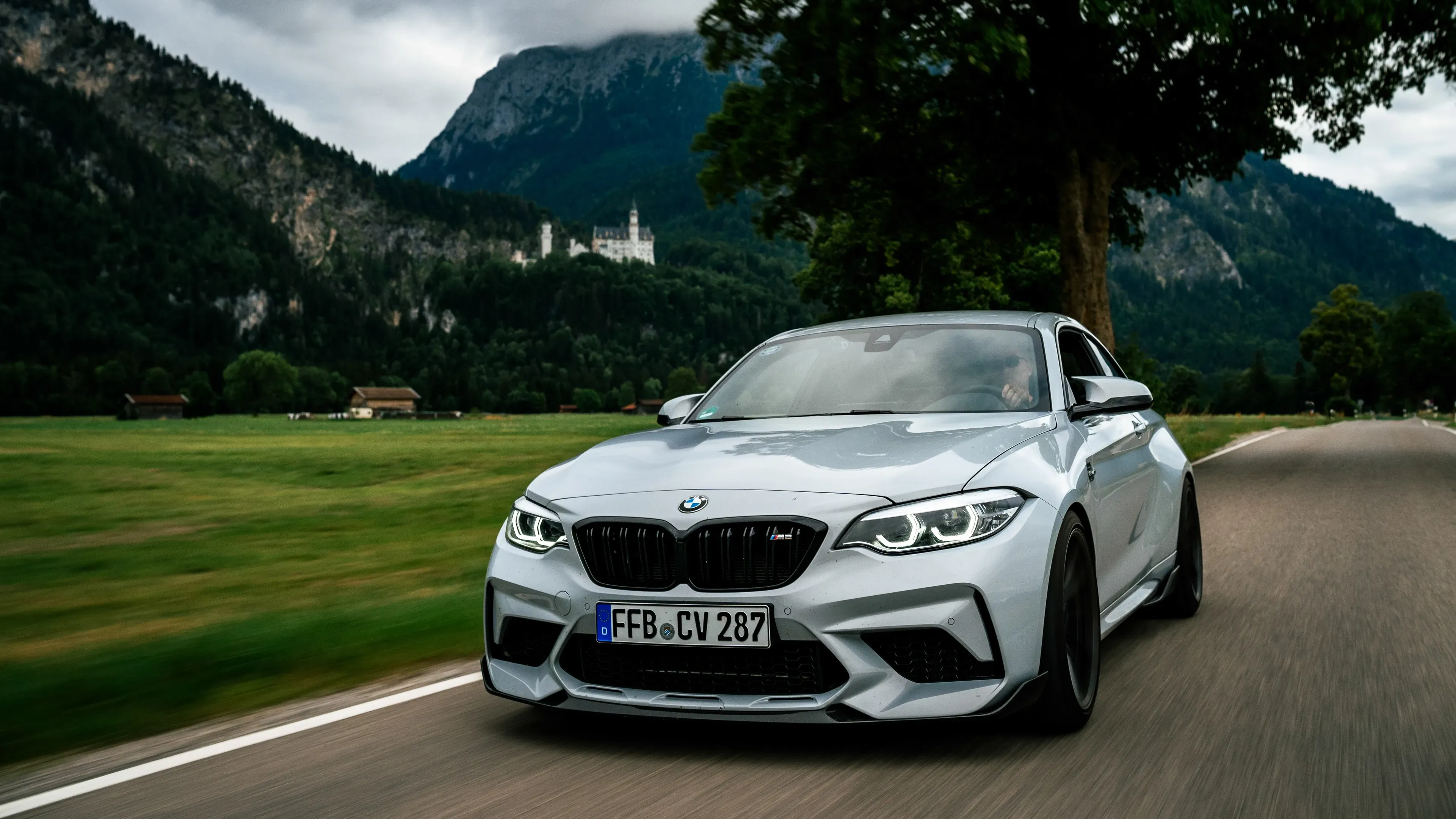 Silberner BMW M2 fährt auf einer Landstraße, im Hintergrund Schloss Neuschwanstein vor bewaldeten Bergen