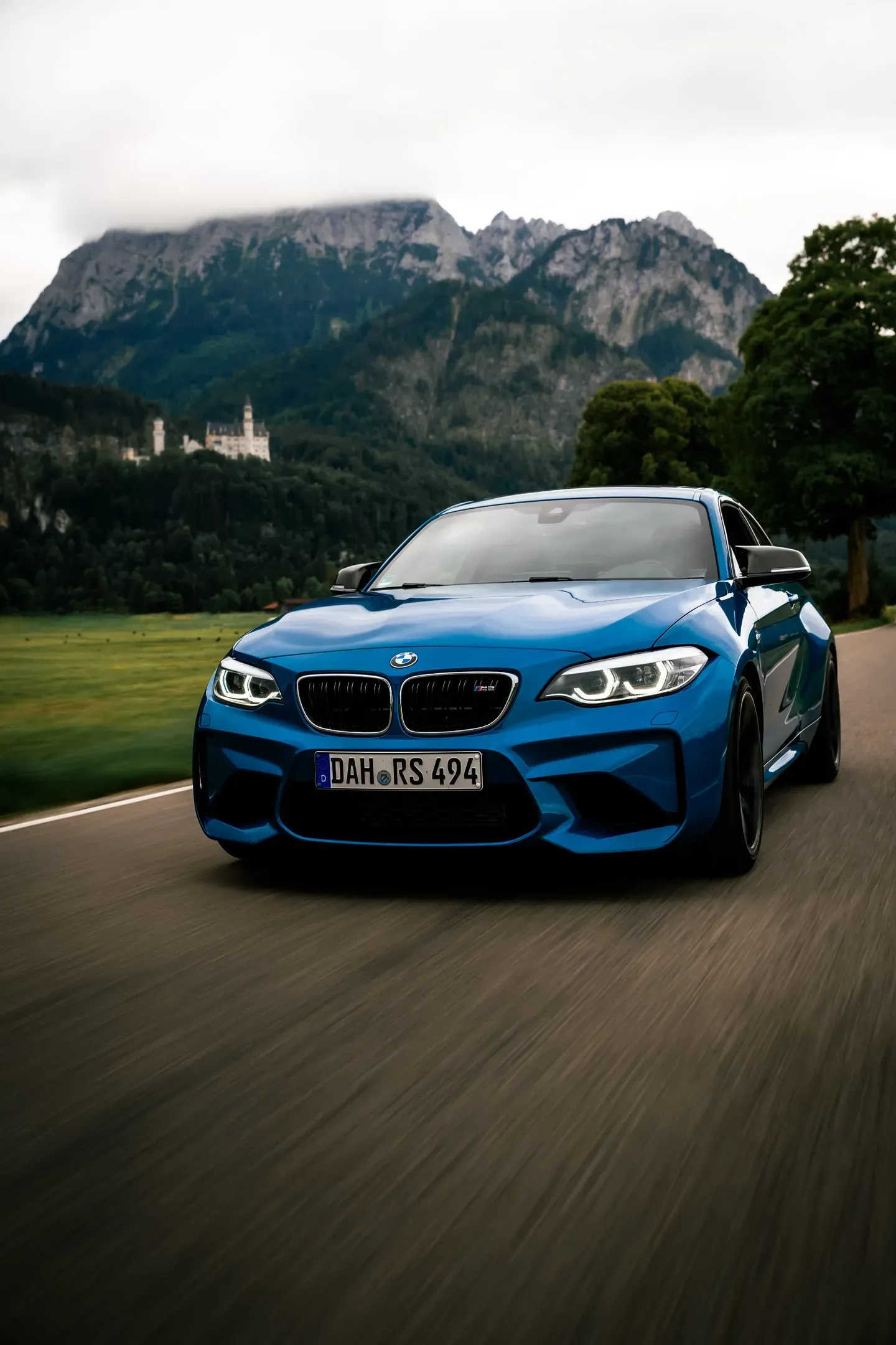 Blauer BMW M2 fährt frontal auf die Kamera zu, dahinter Schloss Neuschwanstein und wolkenverhangene Felsgipfel