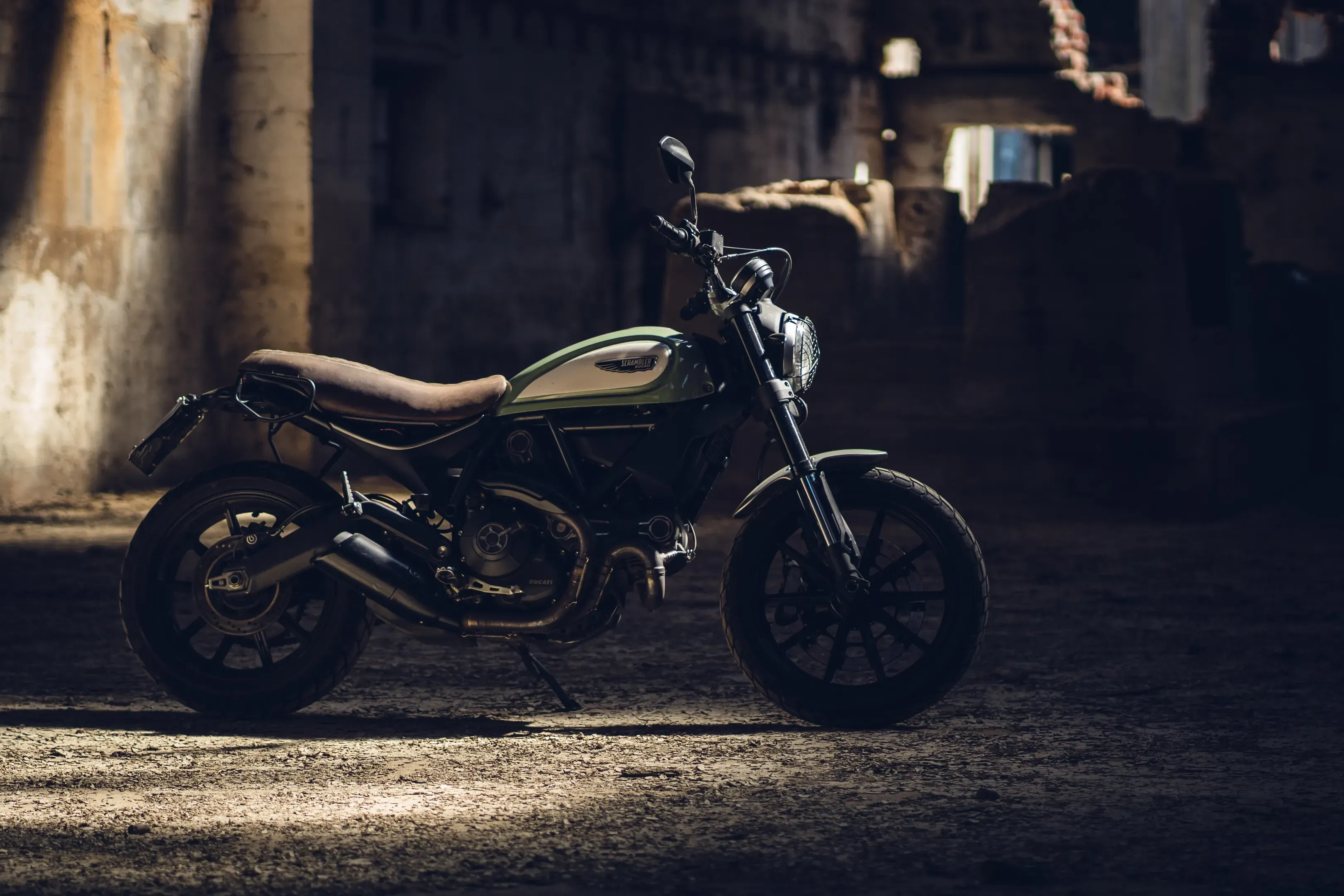 Grün-silbernes Motorrad im Scrambler-Stil mit brauner Sitzbank in einer dunklen, verfallenen Halle mit Lichteinfall