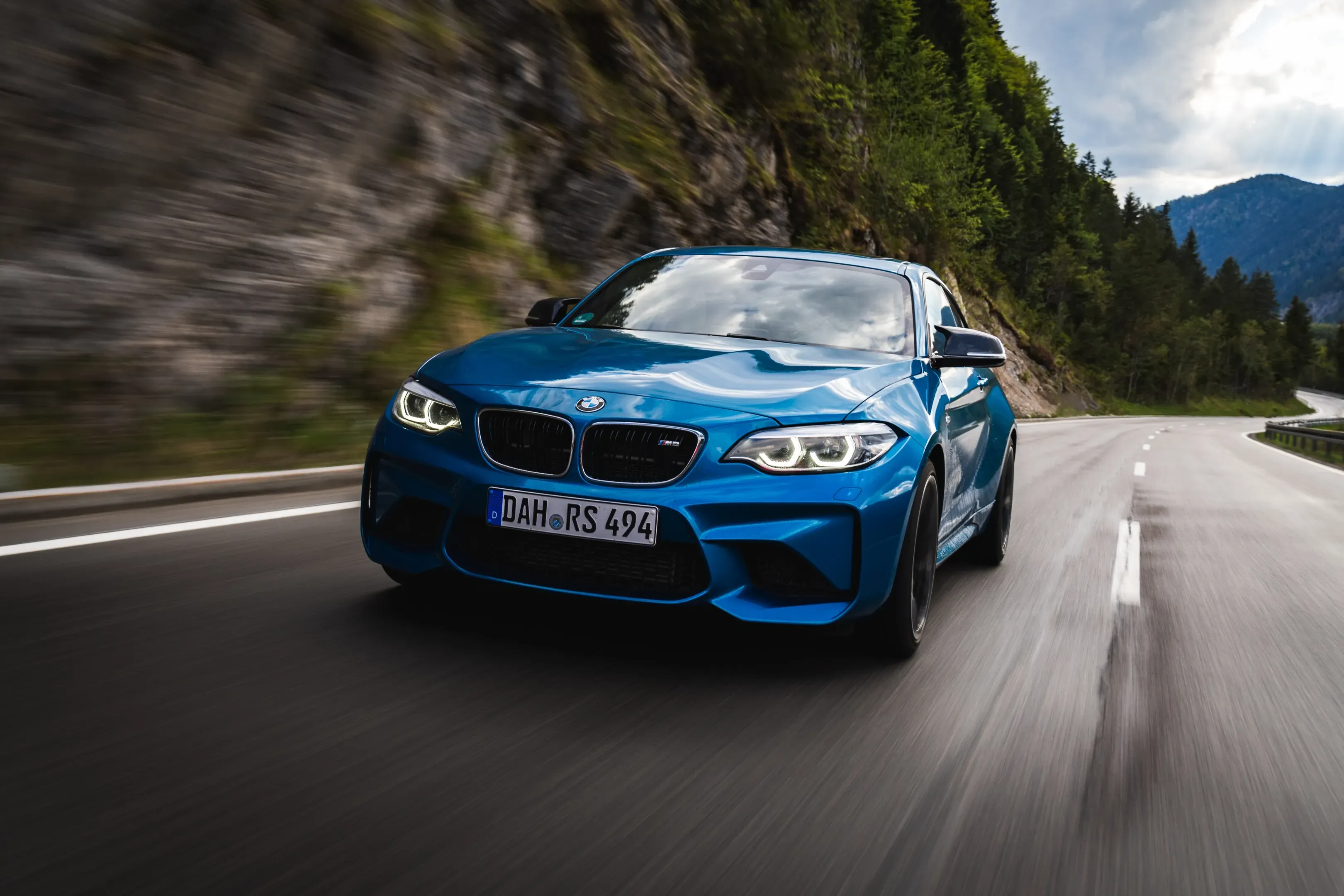 Blauer BMW M2 fährt auf einer nassen Bergstraße durch eine Kurve, dahinter bewaldete Hänge und Felsen