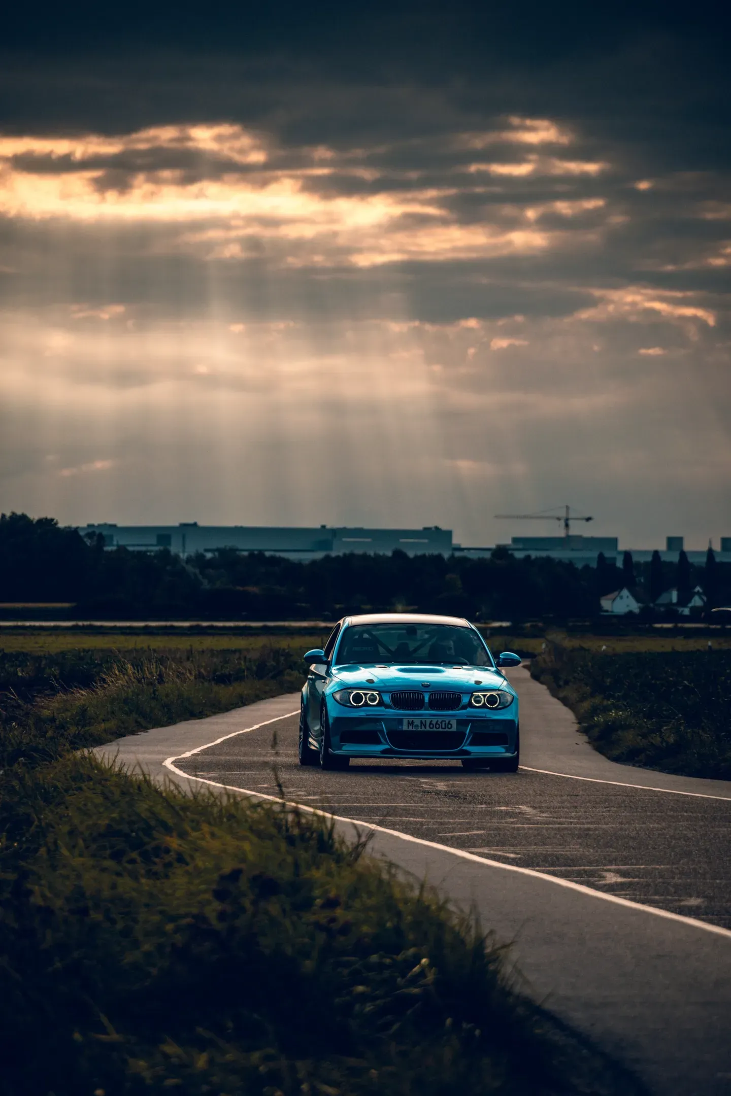 Hellblaues BMW Coupé auf einem schmalen Weg zwischen Feldern, darüber dunkle Wolken mit Sonnenstrahlen
