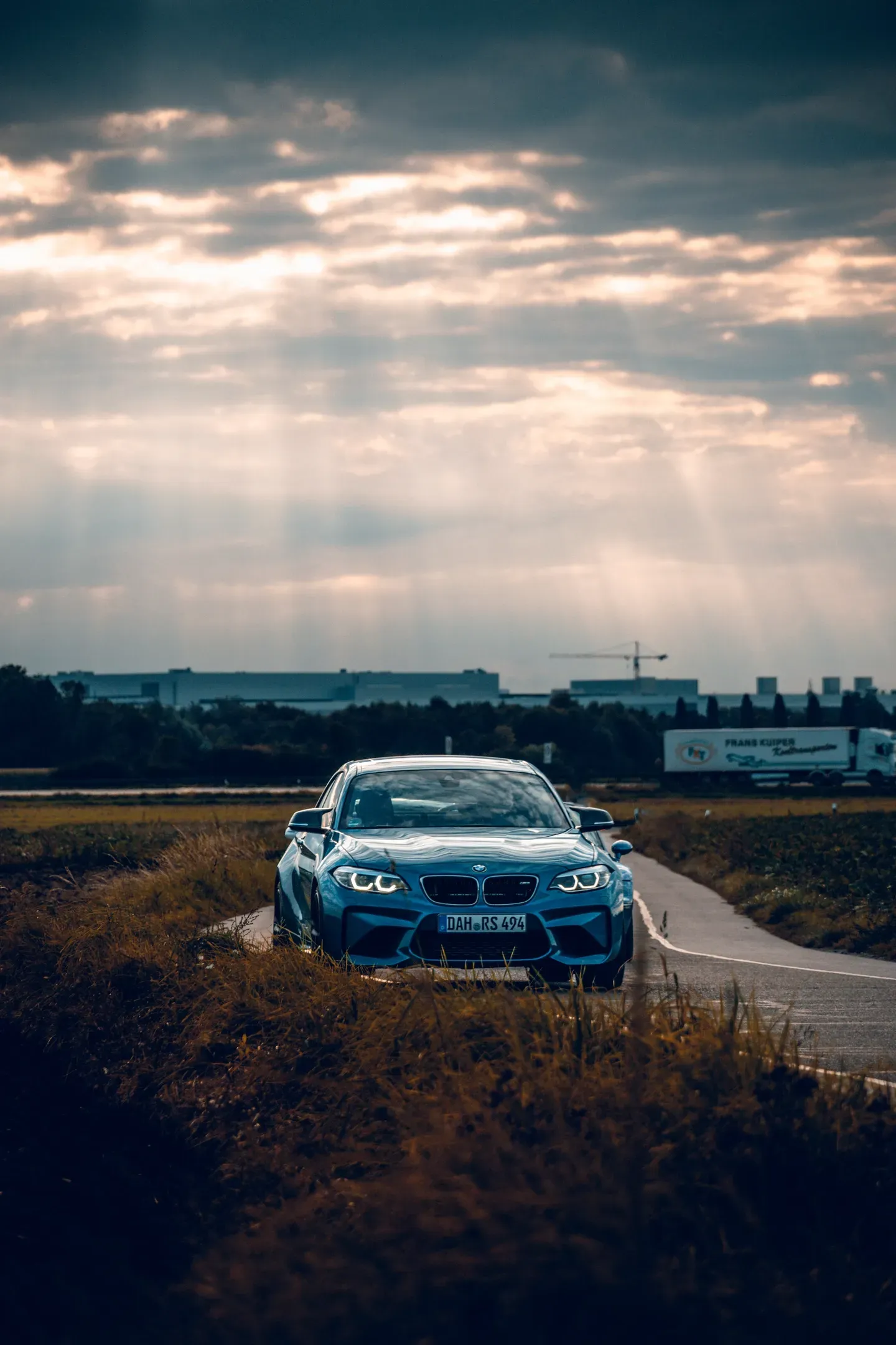 Blauer BMW M2 steht hinter trockenem Gras auf einem Feldweg, Sonnenstrahlen fallen durch dramatische Wolken