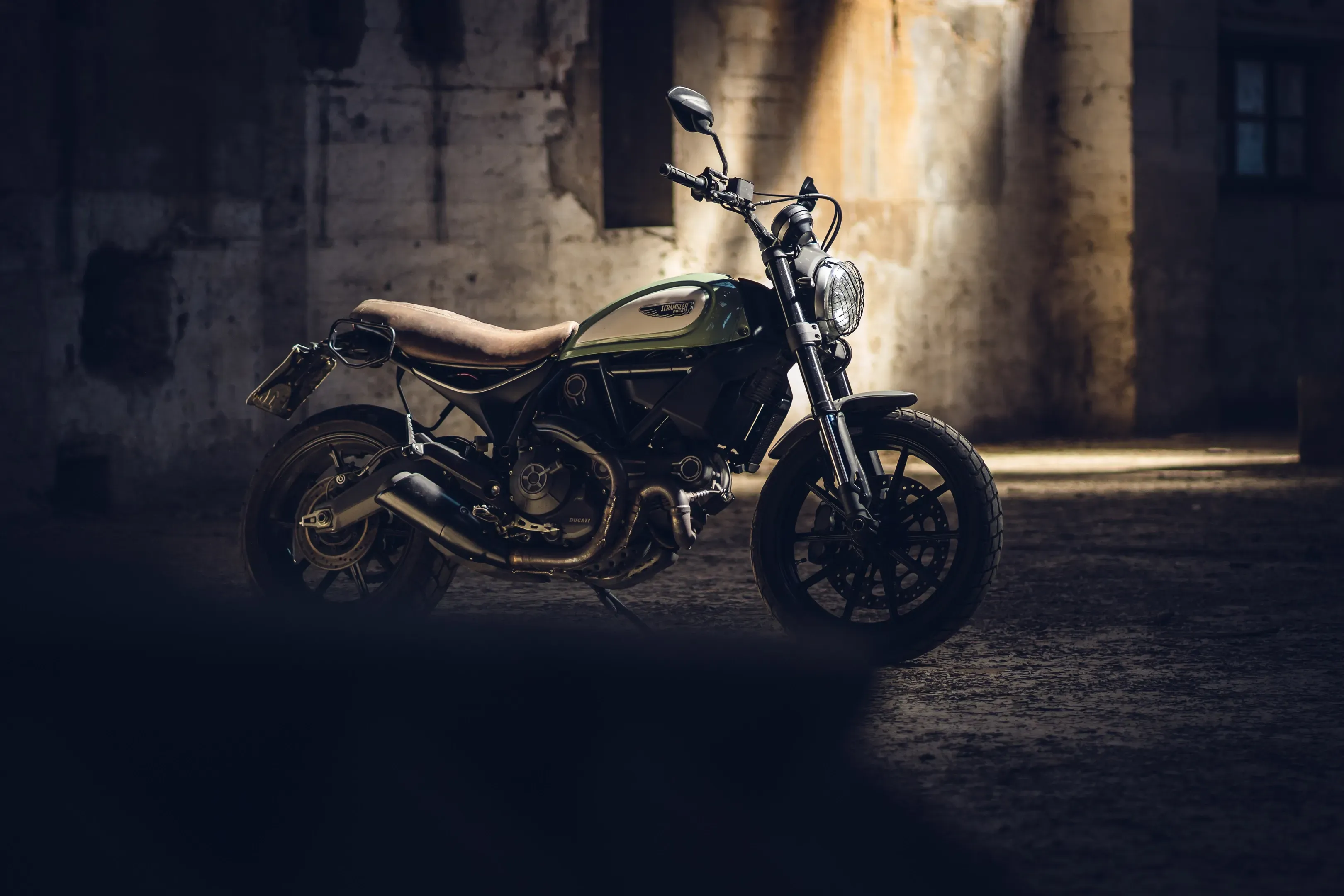 Grün-silbernes Scrambler-Motorrad seitlich in einer verlassenen Halle, ein Lichtstreifen fällt über den Boden