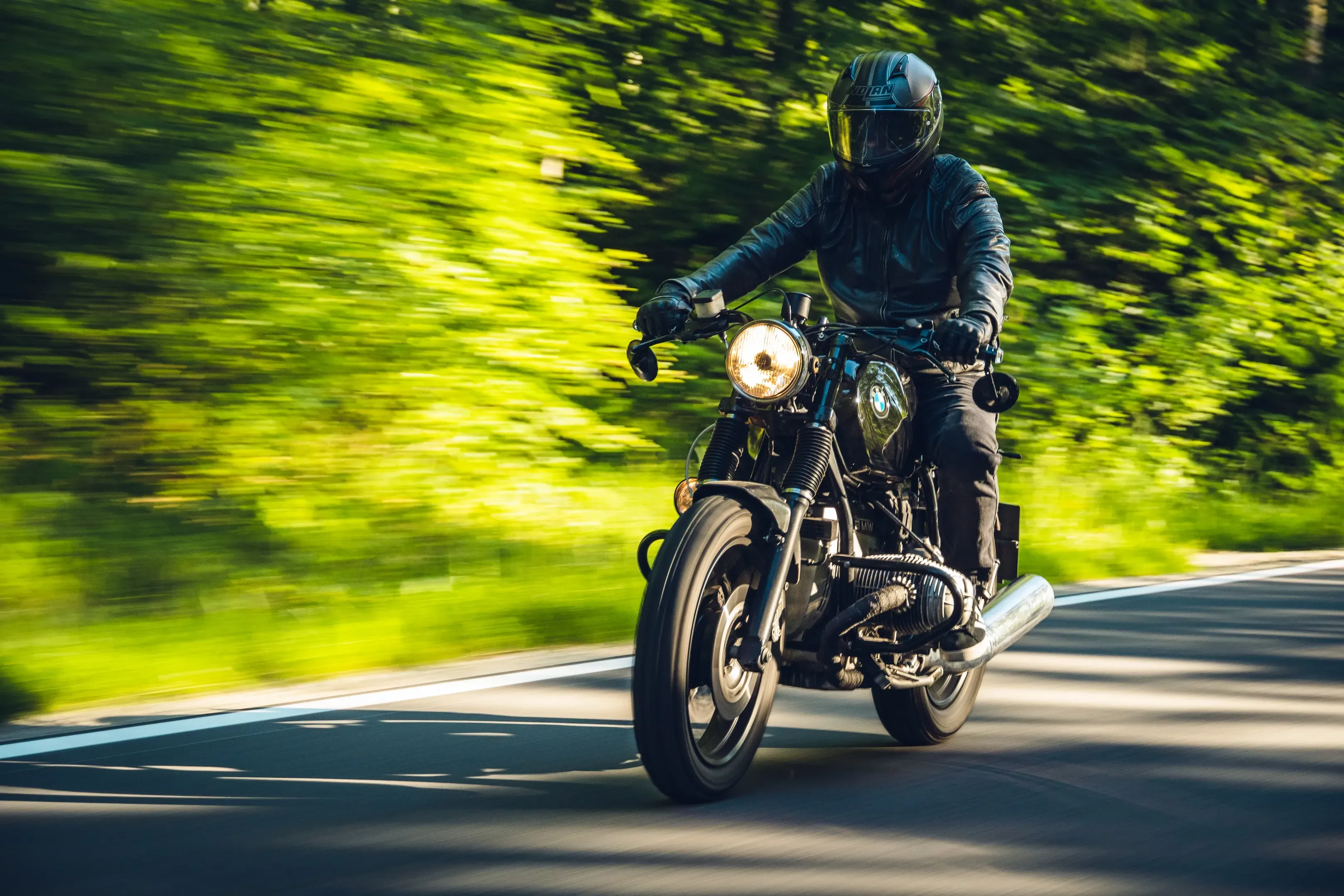 Fahrer in schwarzer Lederjacke auf einem Café Racer mit Boxermotor, frontal auf einer Waldstraße mit Bewegungsunschärfe