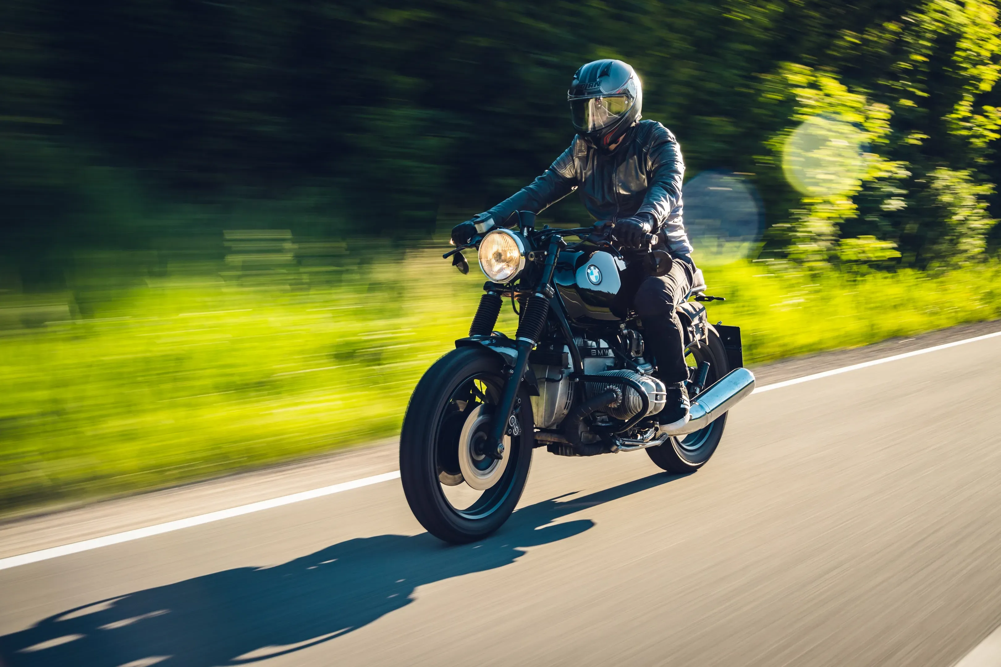 Motorradfahrer auf schwarzem BMW Café Racer im Sonnenlicht, grüner Straßenrand verwischt, Schatten auf dem Asphalt