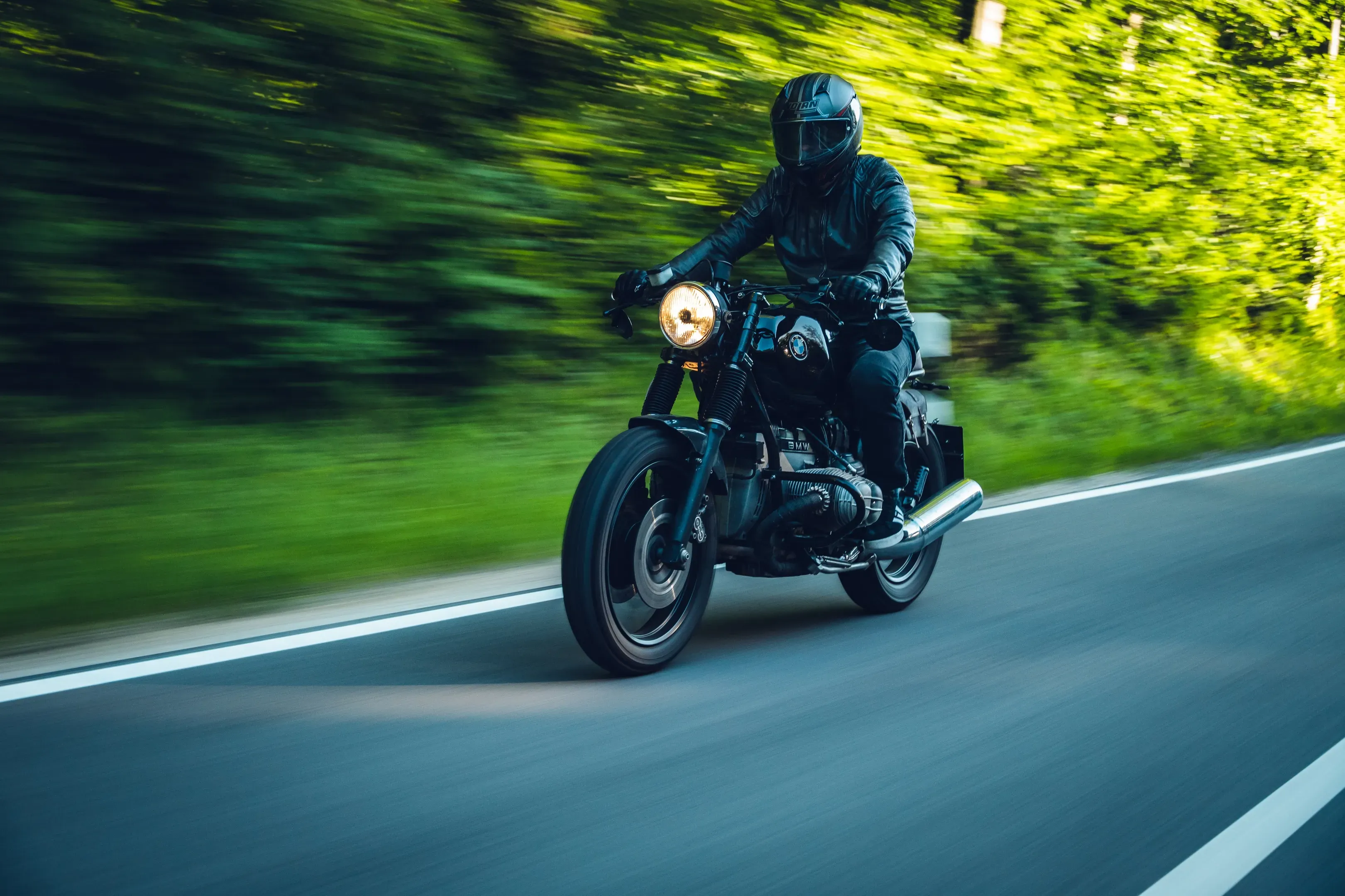 Schwarzer Café Racer mit Fahrer in Lederkleidung auf einer Landstraße, eingeschalteter Rundscheinwerfer, Wald verwischt