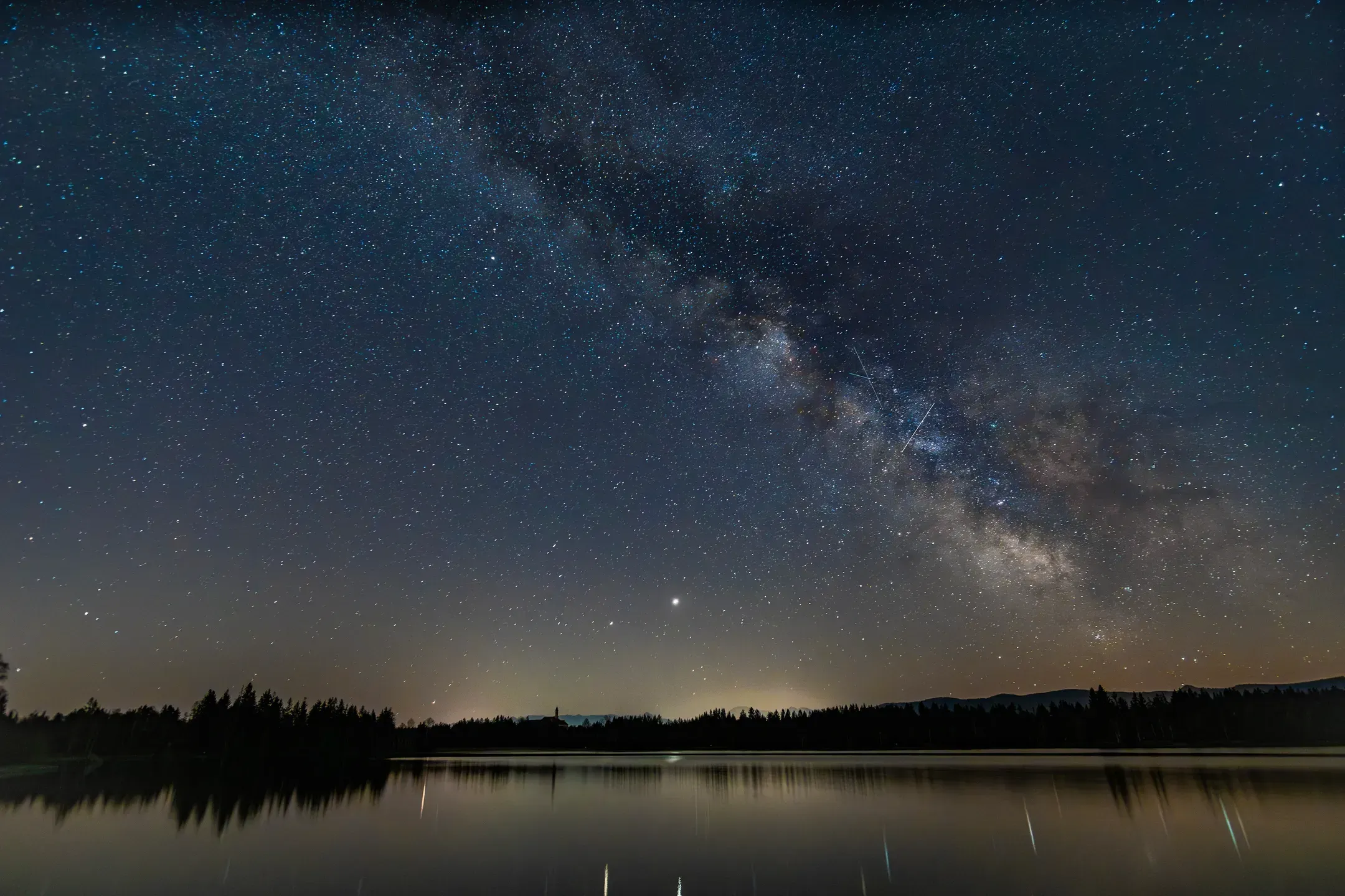 Milchstraße über einem stillen See bei Nacht, Waldsilhouette am Ufer spiegelt sich im Wasser