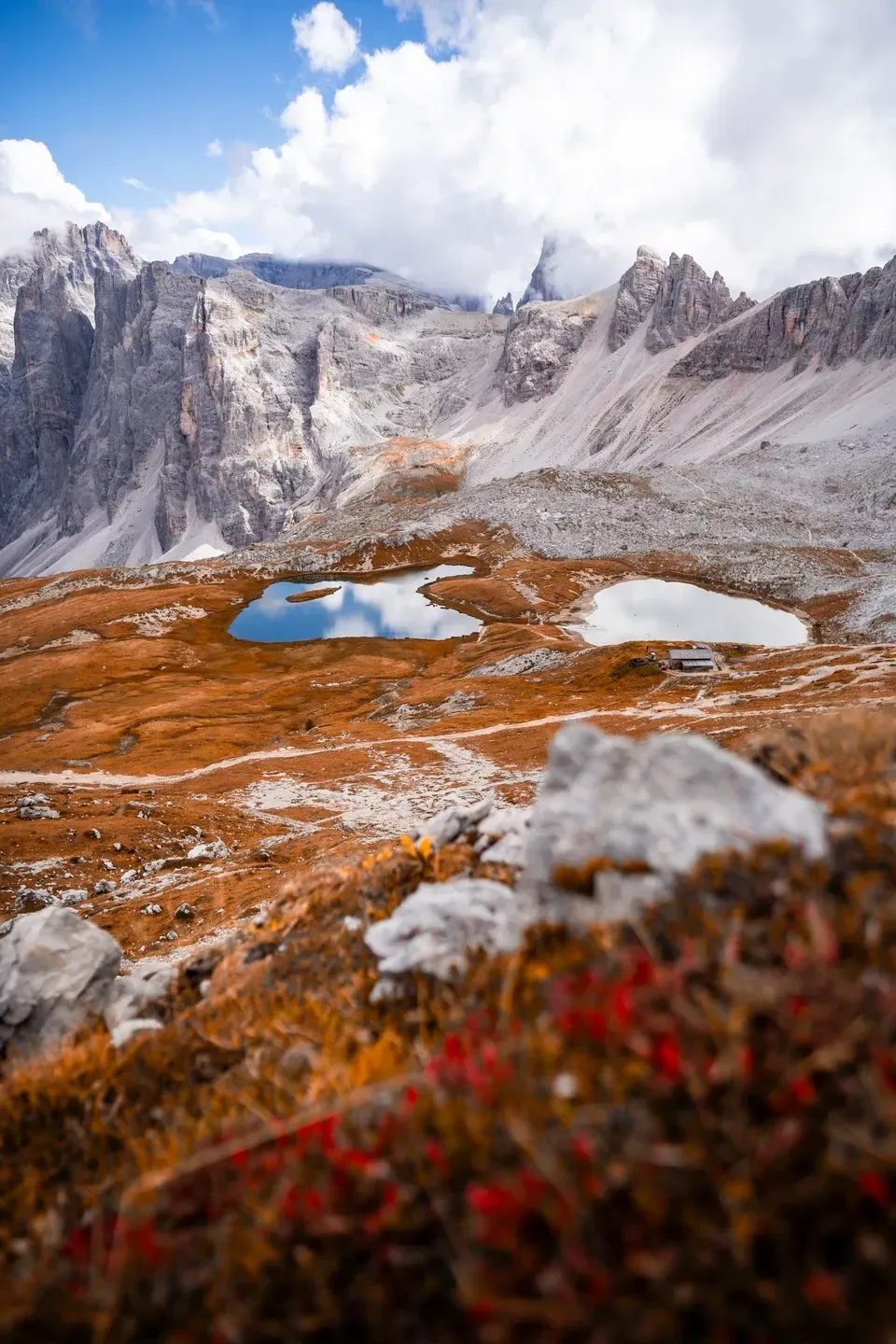 Zwei kleine Bergseen in herbstlich rotbraunem Gelände unter schroffen hellen Felswänden mit Wolken