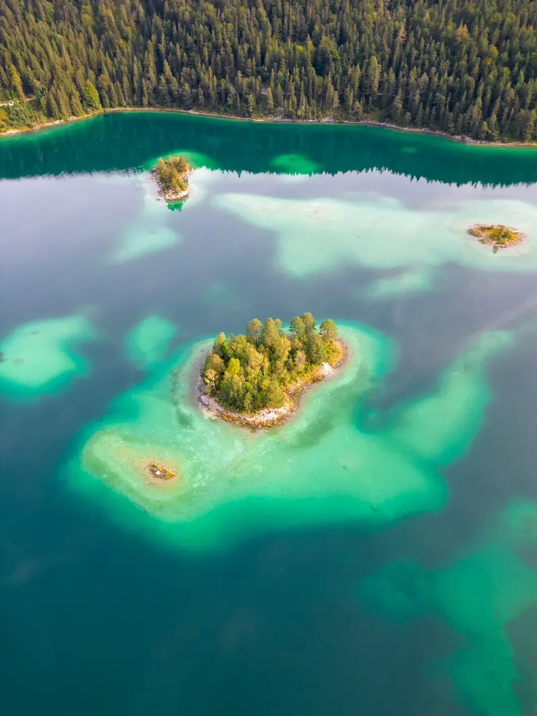 Drohnenaufnahme einer bewaldeten Insel in türkisblauem Bergsee, dahinter dichter Nadelwald am Ufer