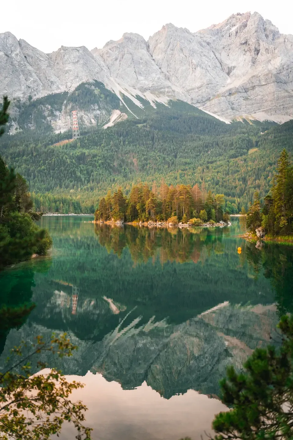 Zugspitzmassiv mit Seilbahnstütze spiegelt sich im Eibsee, davor eine kleine Insel mit Herbstbäumen