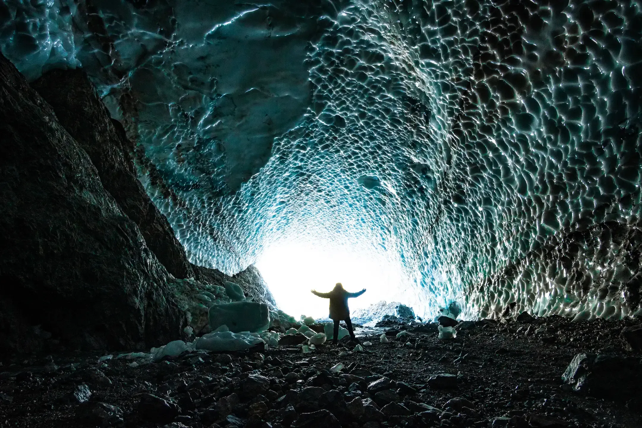 Person steht mit ausgebreiteten Armen am hellen Ausgang einer Eishöhle mit wabenartig geformter Decke