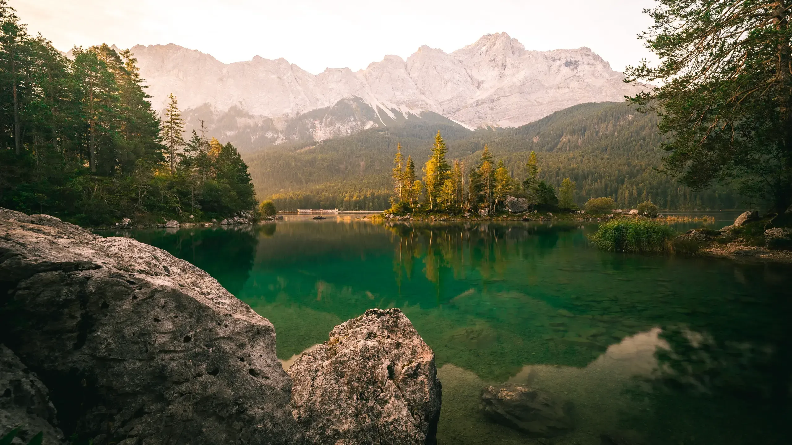 Eibsee mit klarem grünem Wasser und Felsbrocken im Vordergrund, dahinter das Zugspitzmassiv im Morgenlicht