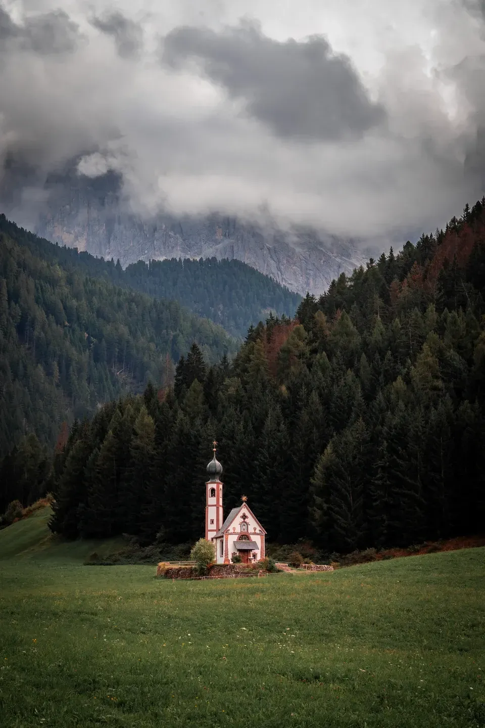 Kirchlein St. Johann in Ranui auf einer Wiese vor dunklem Nadelwald, darüber Wolken an den Felswänden
