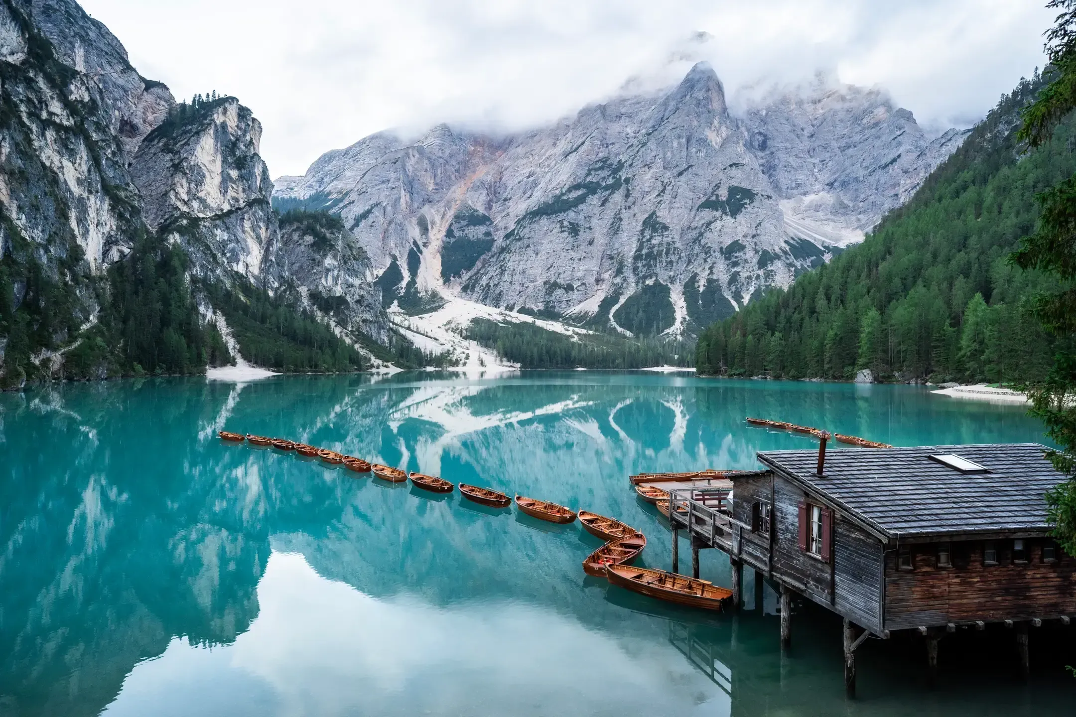 Pragser Wildsee mit türkisem Wasser, Holzbootshaus und einer Reihe Ruderboote vor wolkenverhangenen Felswänden