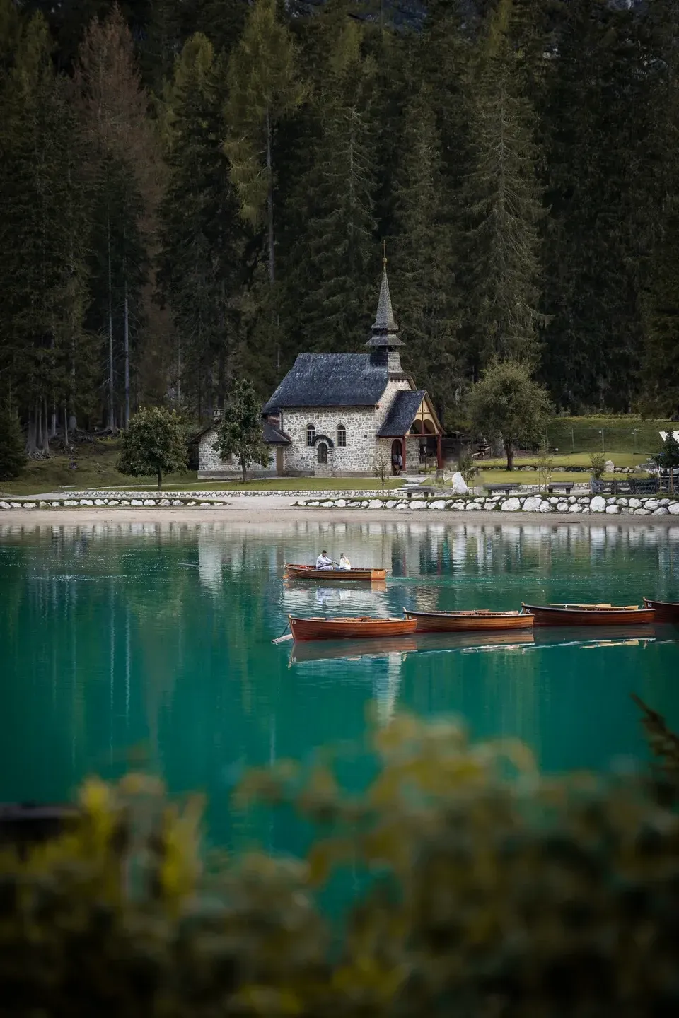 Kleine Steinkapelle am Ufer eines türkisen Bergsees vor dunklem Nadelwald, davor Ruderboote auf dem Wasser