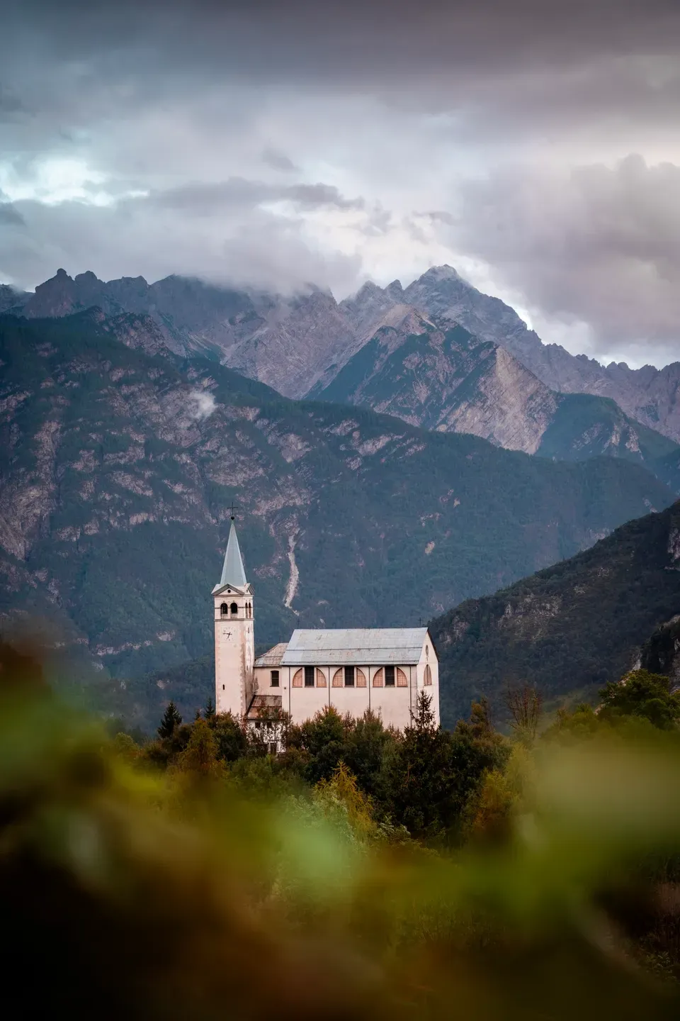 Weiße Kirche mit spitzem Glockenturm vor wolkenverhangenen Bergen, im Vordergrund unscharfes Grün