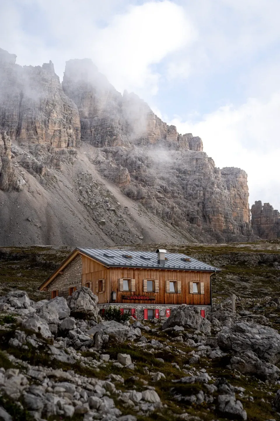 Berghütte aus Holz mit roten Fensterläden zwischen Felsblöcken, dahinter Nebel an steilen Dolomitenwänden
