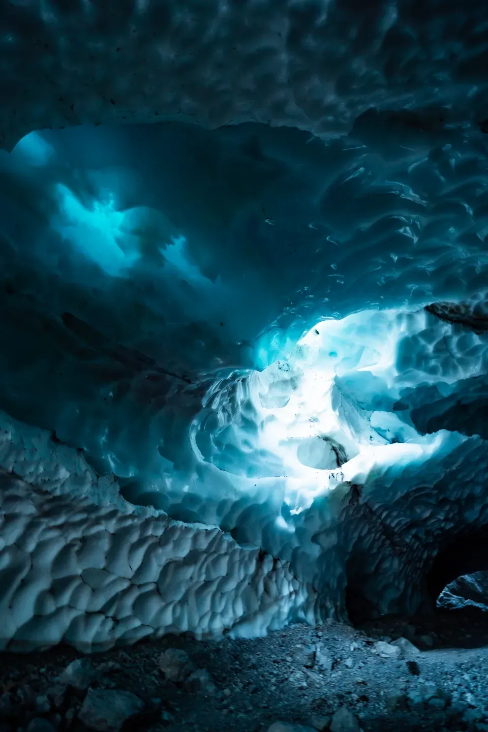 Blick in eine Eishöhle mit gewellten, blau schimmernden Wänden, durch eine Öffnung fällt helles Licht