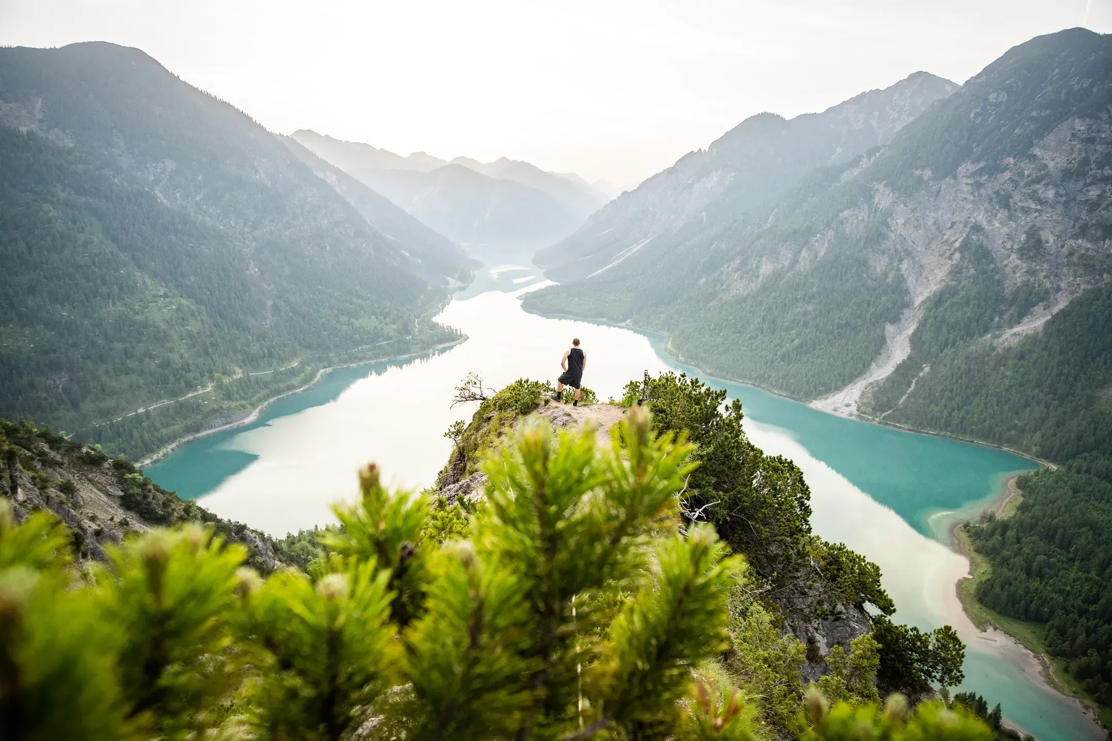 Wanderer steht auf einem Felsvorsprung über einem langgezogenen türkisen Bergsee zwischen bewaldeten Hängen