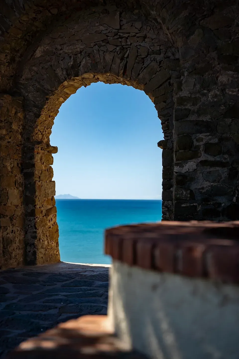 Steinerner Torbogen gibt den Blick auf das tiefblaue Meer frei, in der Ferne eine Insel im Dunst