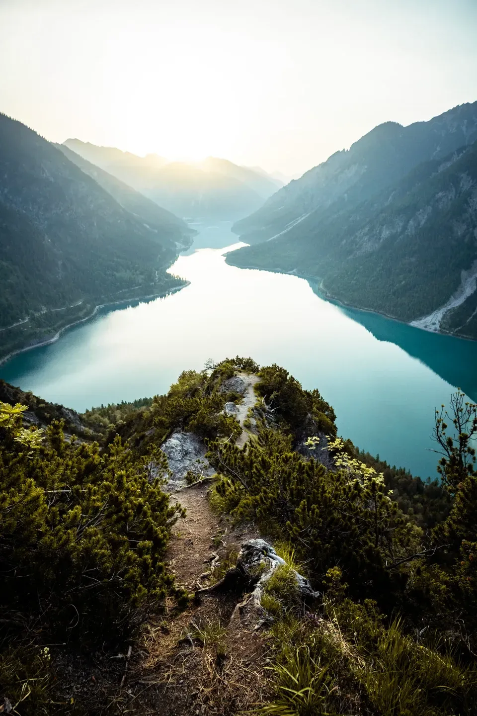 Schmaler Pfad führt zu einem Felsvorsprung über einem langen türkisen Bergsee im Gegenlicht