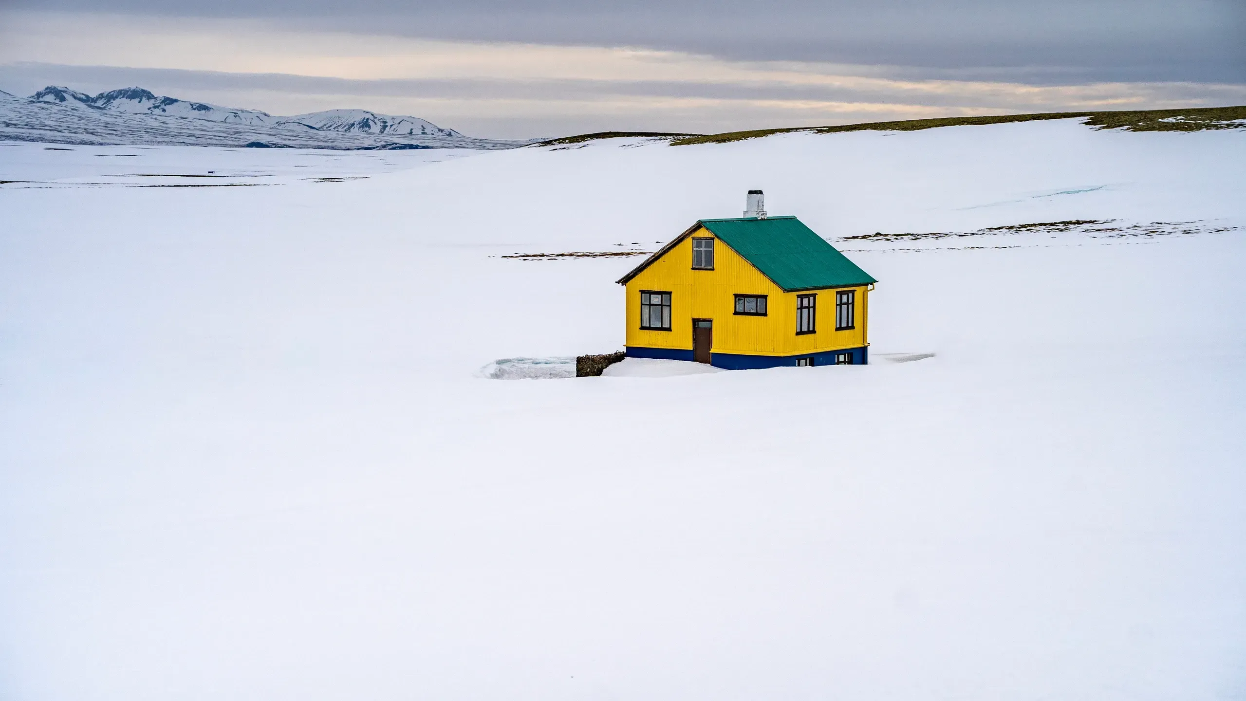 Einzelnes gelbes Haus mit grünem Dach in einer weiten verschneiten Ebene, dahinter flache Berge