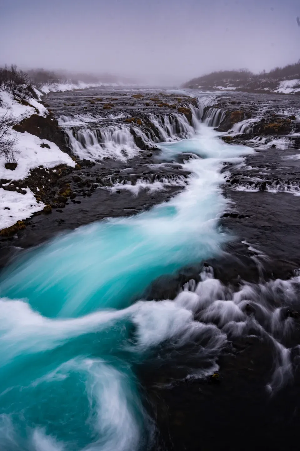 Türkises Wasser strömt in Langzeitbelichtung über dunkle Felsstufen eines Wasserfalls, Ufer mit Schnee