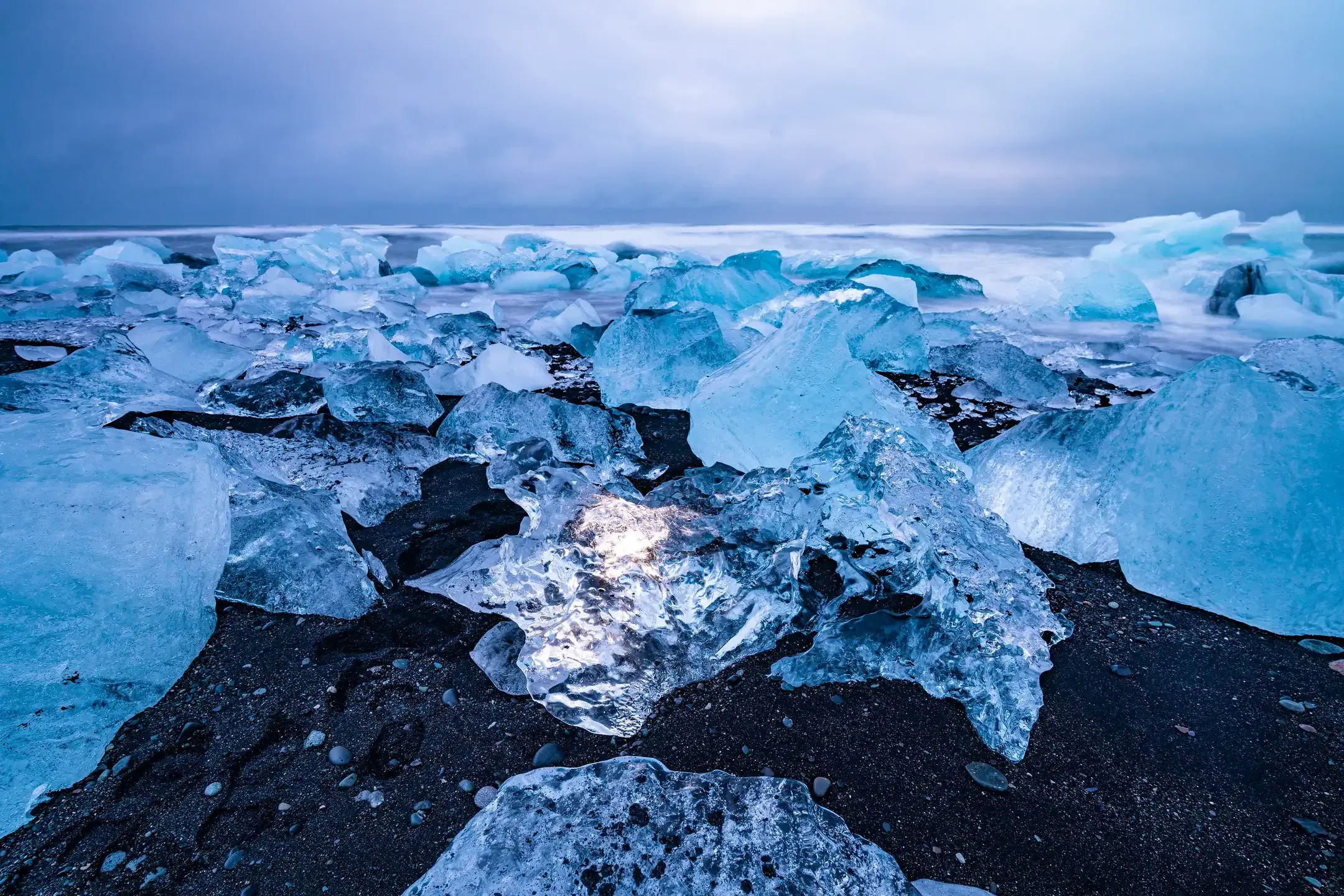 Leuchtende Eisbrocken auf schwarzem Sandstrand in der blauen Stunde, dahinter die Brandung