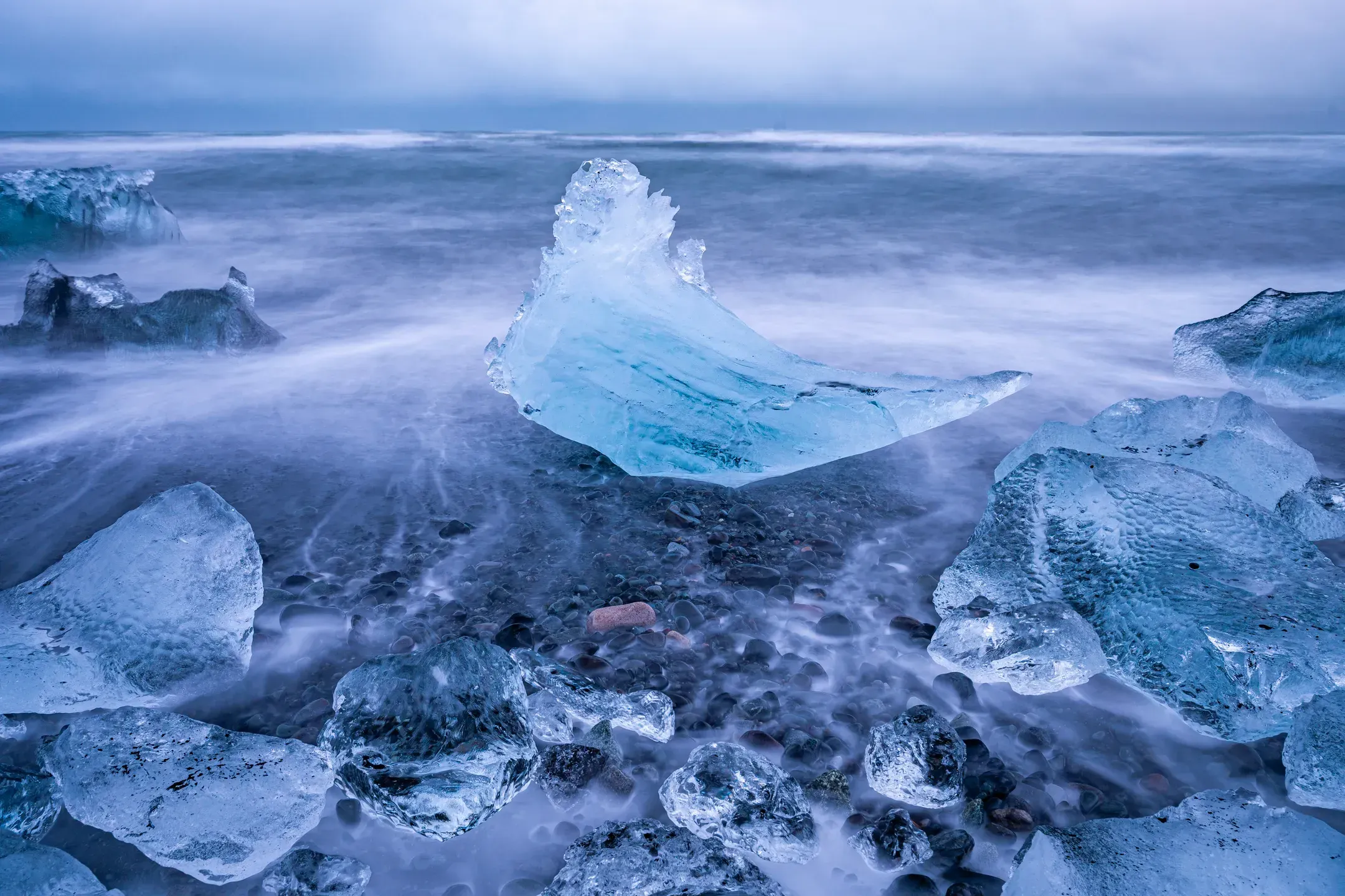 Spitz geformter Eisblock in der Brandung, davor kleinere Eisstücke und dunkle Kiesel am Strand