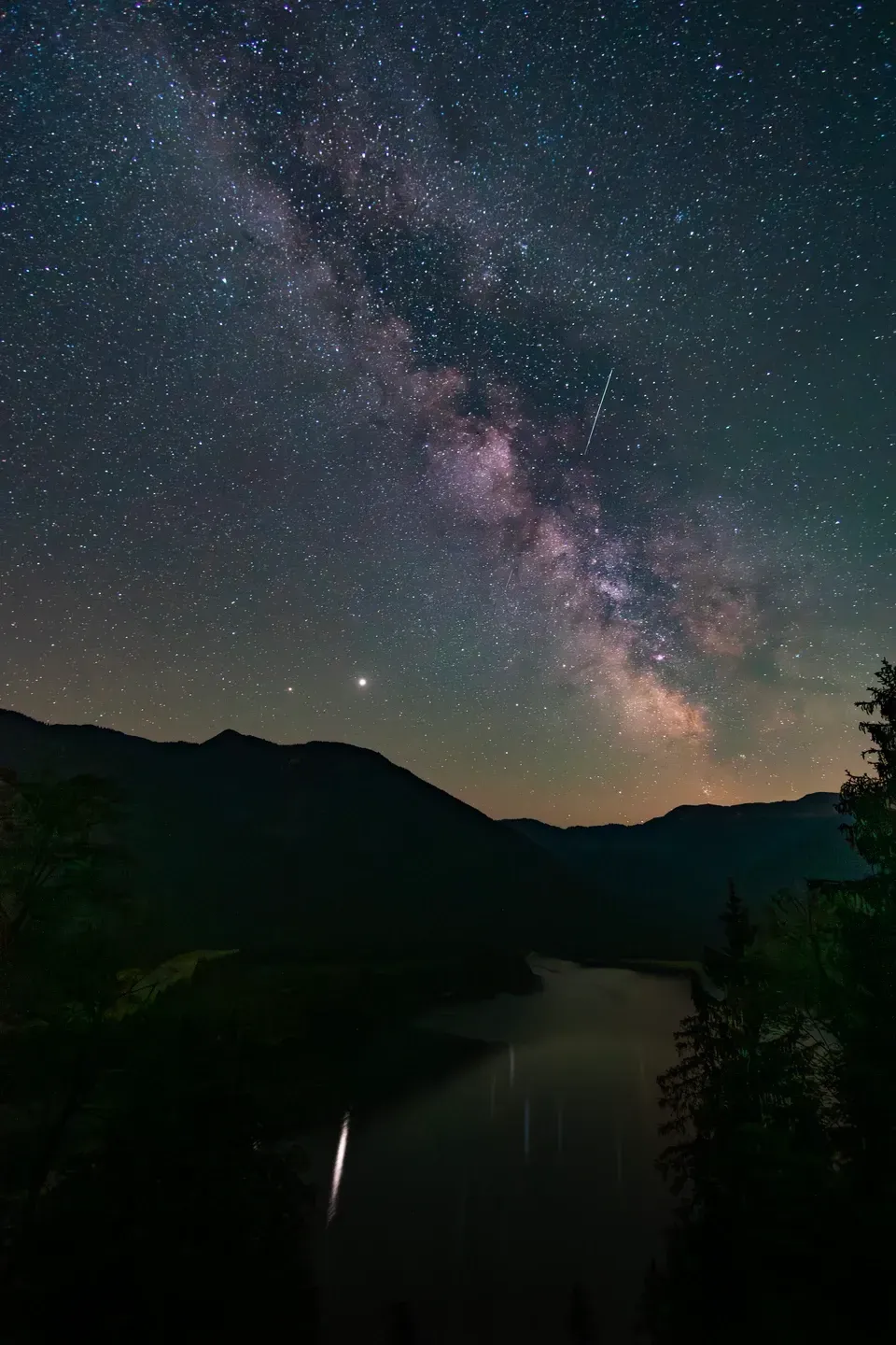 Milchstraße und Sternschnuppe über einem dunklen Bergsee, ein heller Planet über den Hügeln