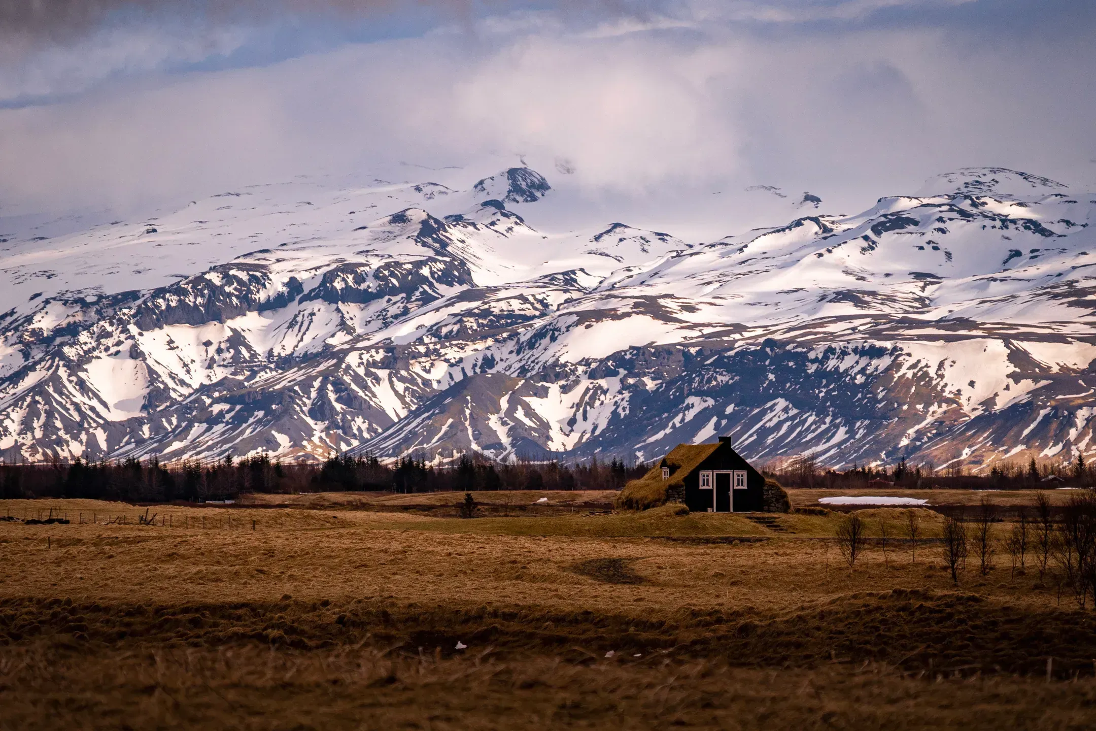 Schwarzes Torfhaus mit Grasdach auf einer braunen Wiese, dahinter schneebedeckte Berge