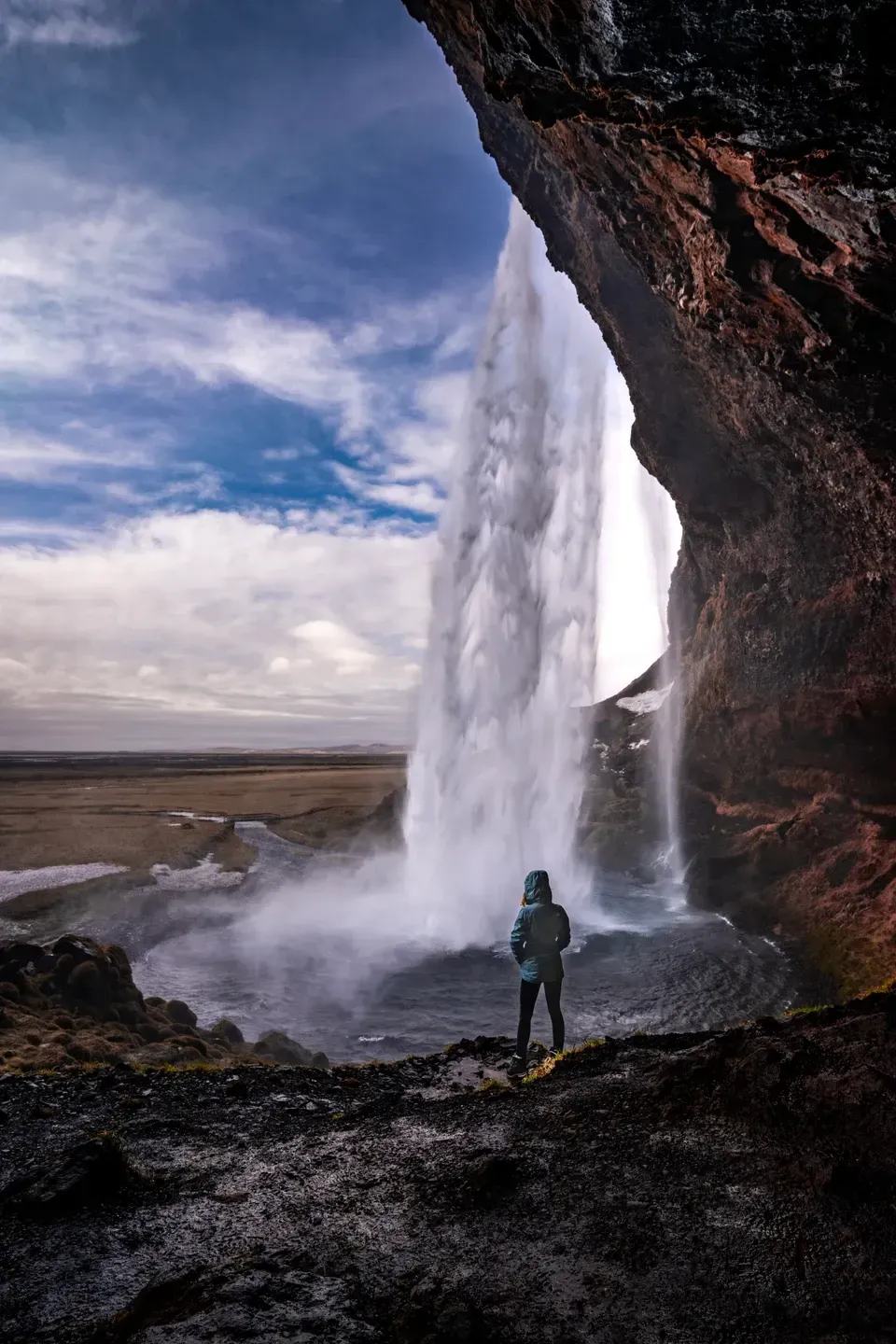 Person in Regenjacke steht hinter einem Wasserfall unter einem Felsüberhang und blickt ins Freie