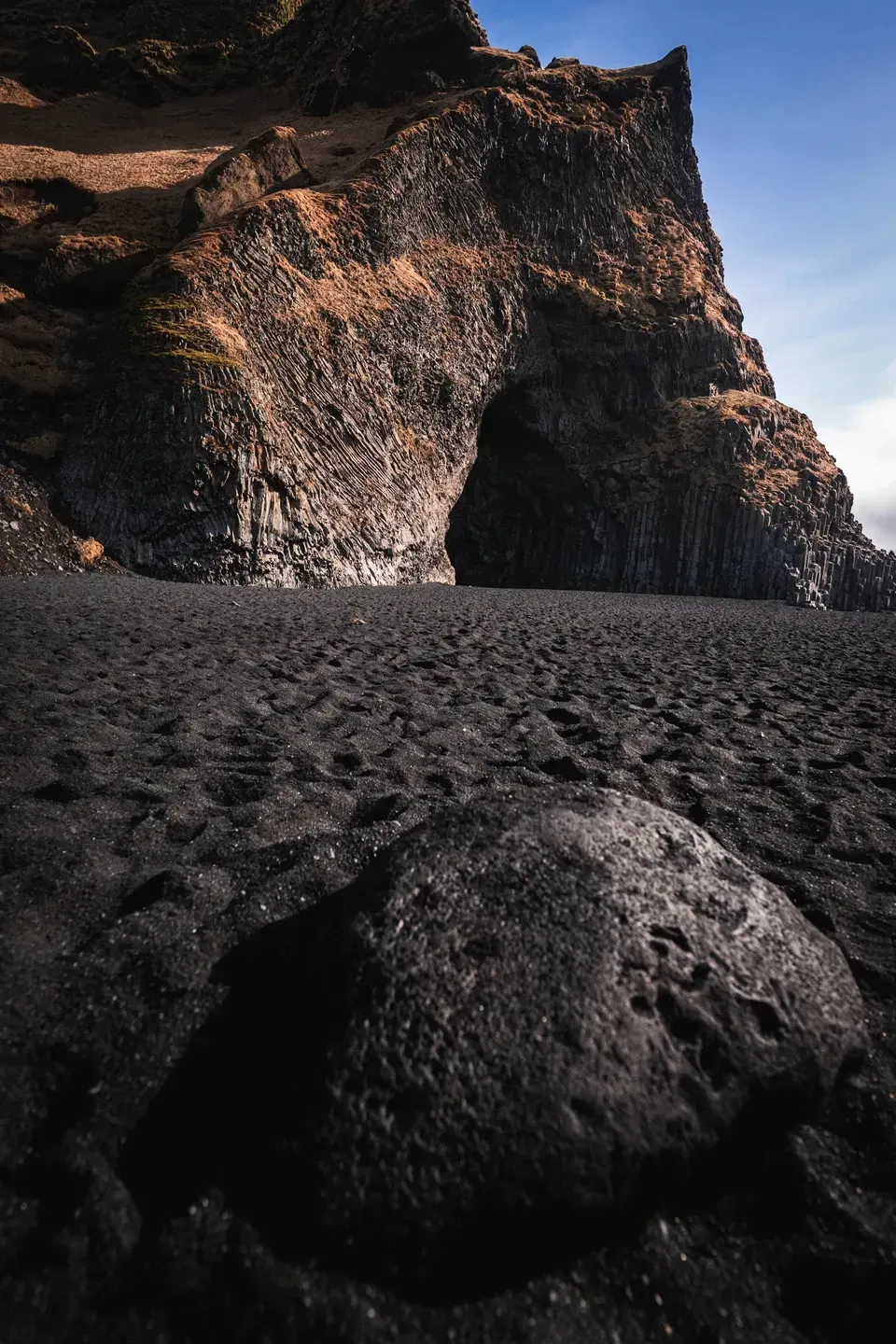 Großer Lavastein auf schwarzem Sand vor einer Basaltklippe mit Höhlenöffnung