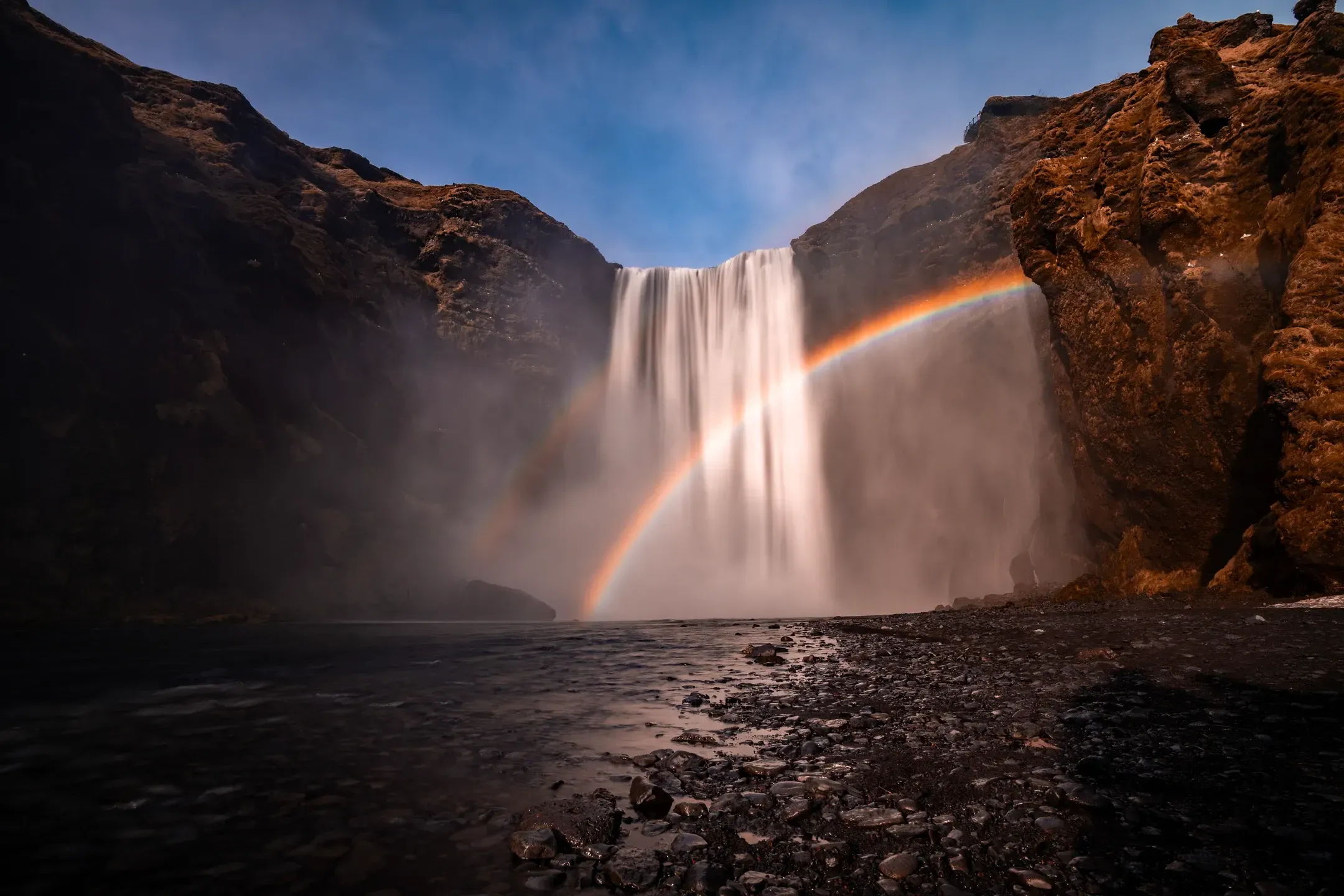 Breiter Wasserfall stürzt über eine Felskante, in der Gischt bildet sich ein Regenbogen