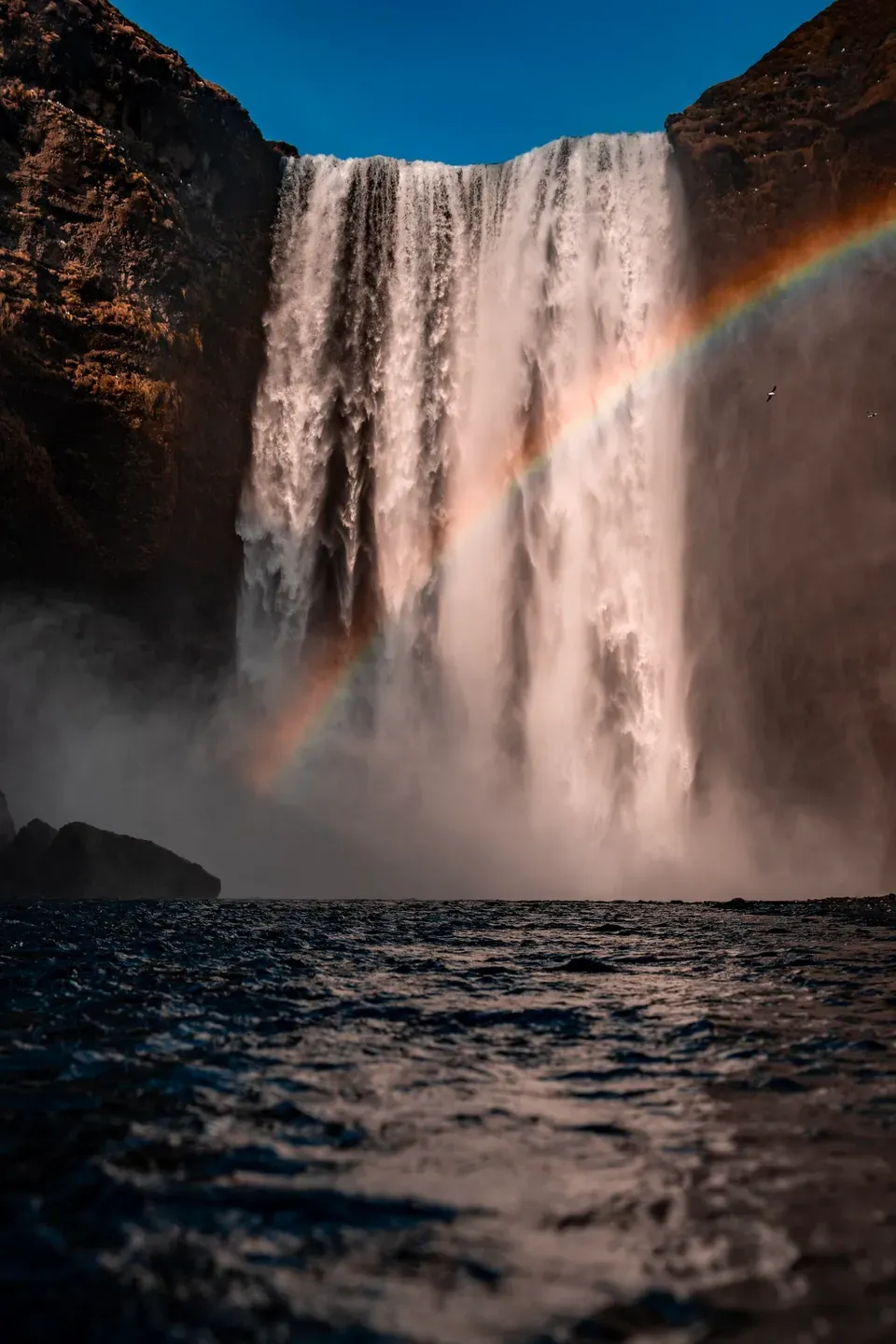 Wasserfall von unten im Hochformat, Regenbogen in der Gischt und dunkles Wasser im Vordergrund