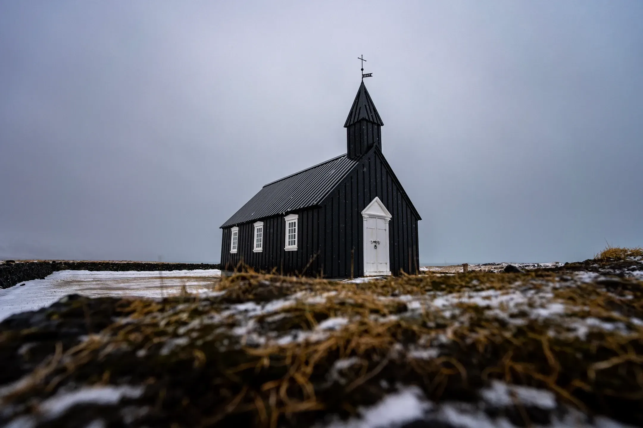 Schwarze Holzkirche mit weißer Tür auf winterlicher Wiese unter grauem Himmel