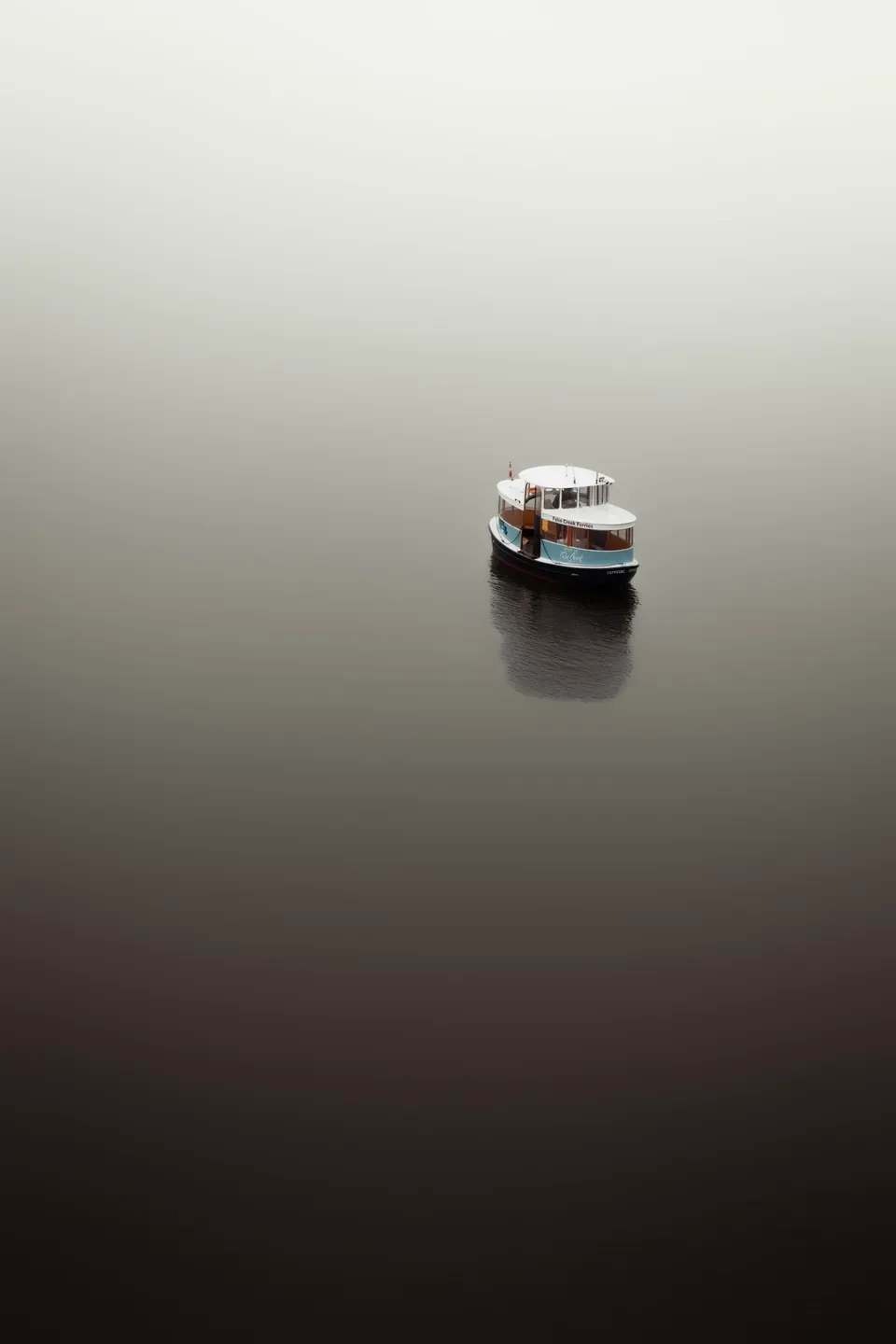 Kleines Fährschiff auf spiegelglattem Wasser von oben, minimalistisch in grauem Nebel