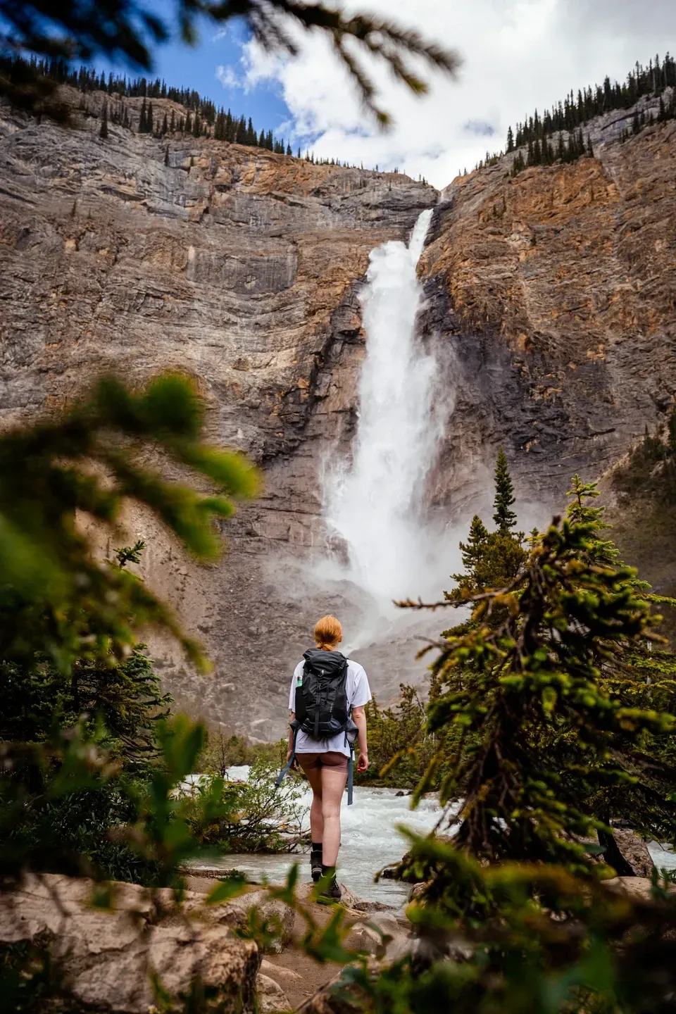 Wanderin mit Rucksack steht vor einem hohen Wasserfall, der über eine steile Felswand ins Tal stürzt