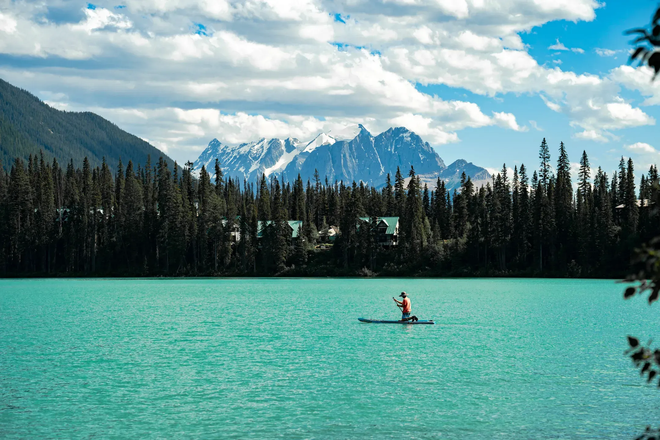 Stand-up-Paddler kniet auf seinem Board auf einem türkisen Bergsee, dahinter Nadelwald und verschneite Gipfel