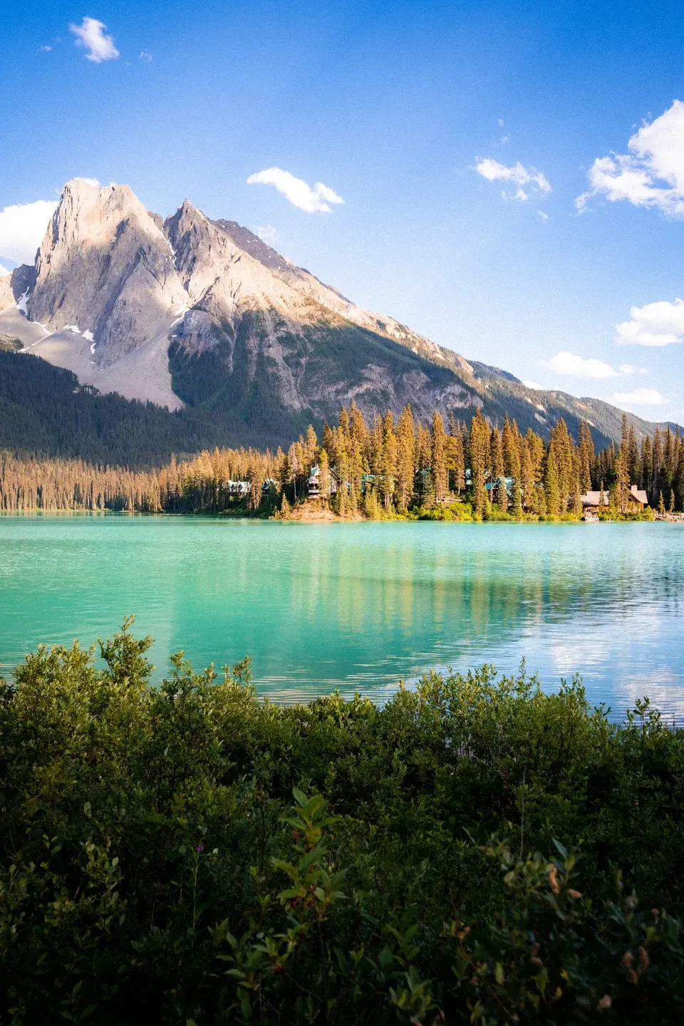 Türkiser Bergsee mit Lodges am bewaldeten Ufer und hellem Felsgipfel im warmen Abendlicht, Büsche im Vordergrund