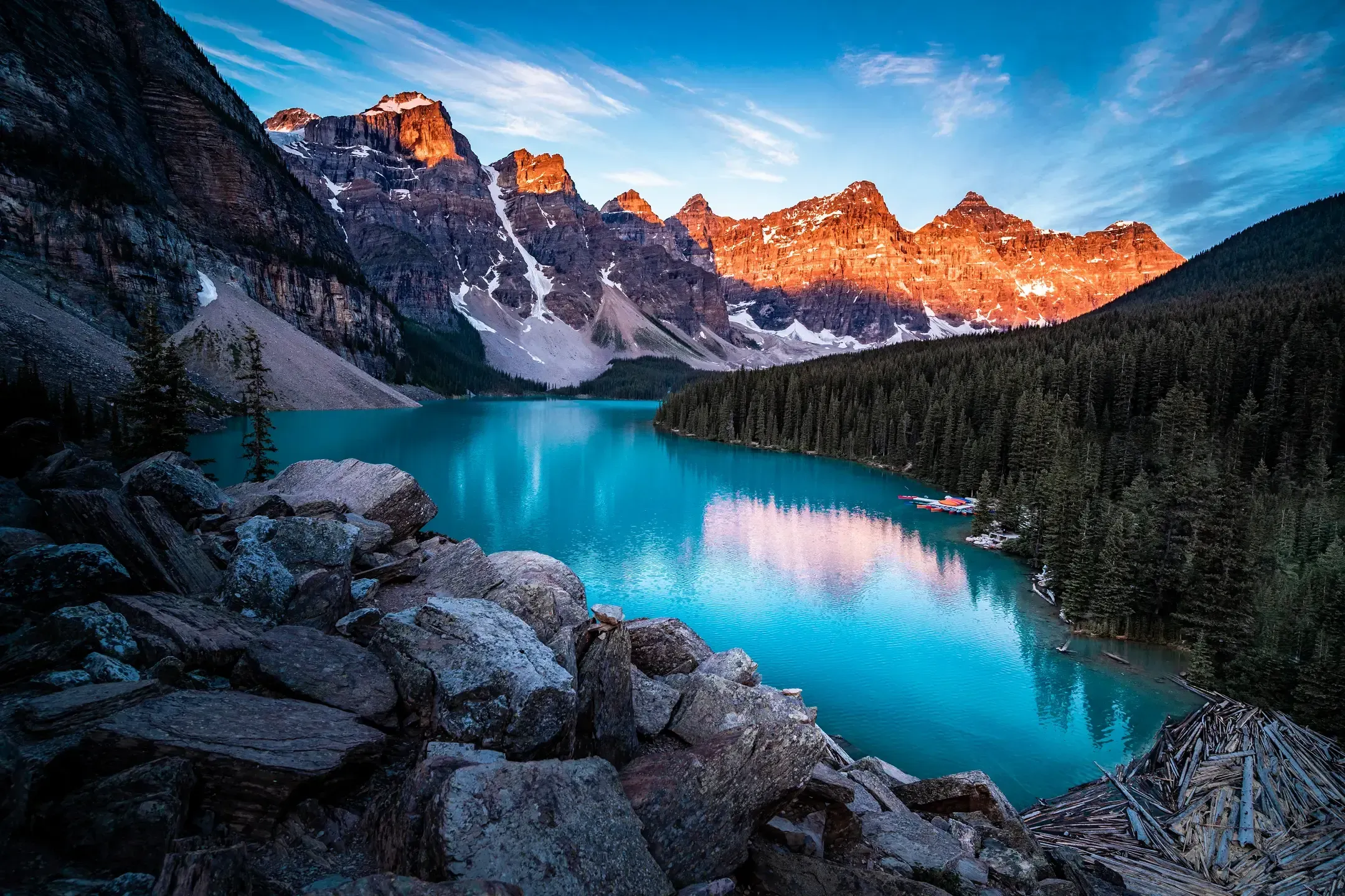 Moraine Lake mit türkisem Wasser, die Gipfel dahinter leuchten im Alpenglühen, Felsblöcke im Vordergrund