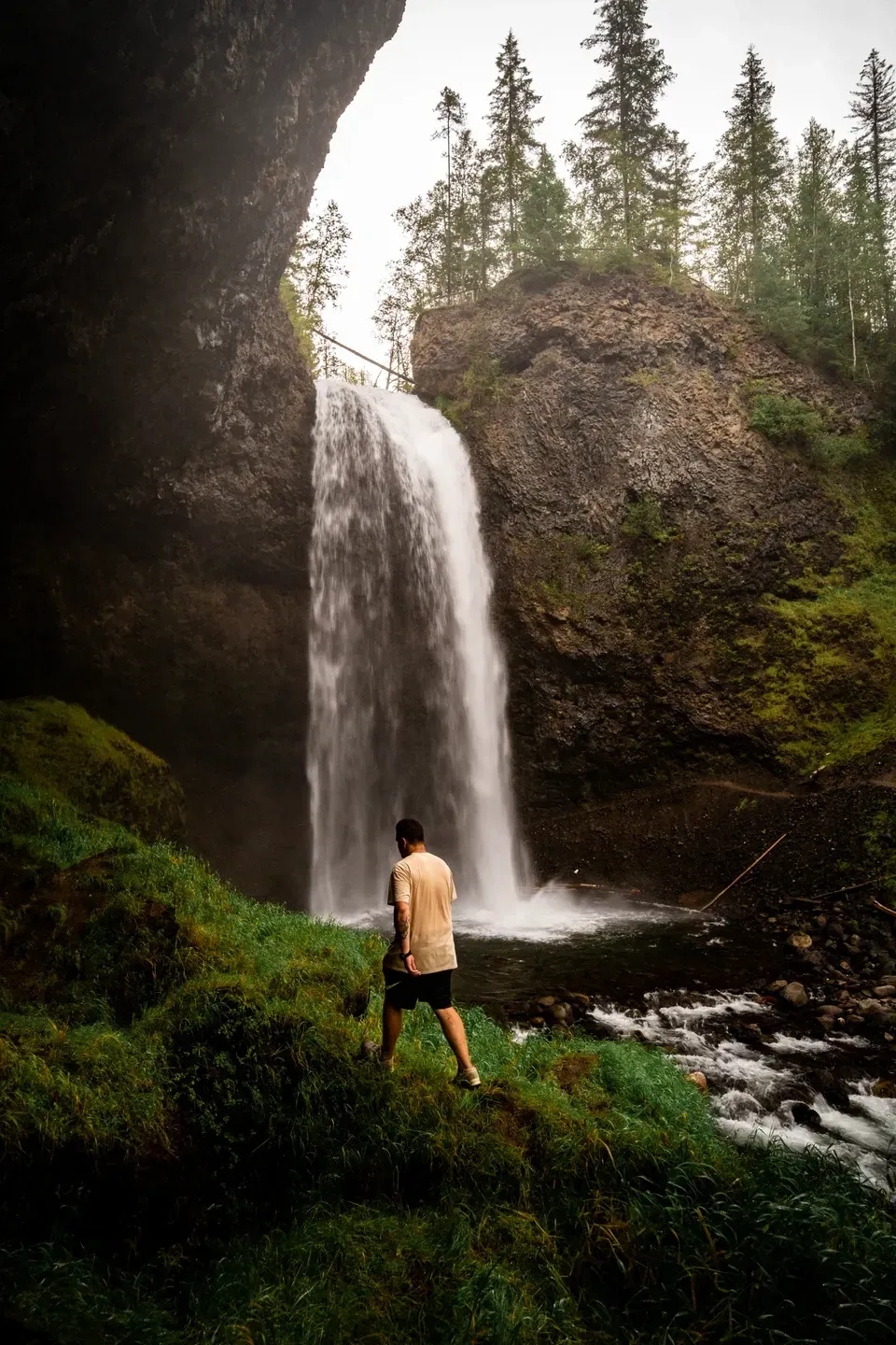 Mann in hellem T-Shirt steht auf einer grünen Wiese vor einem Wasserfall, der über eine Felskante ins Becken fällt