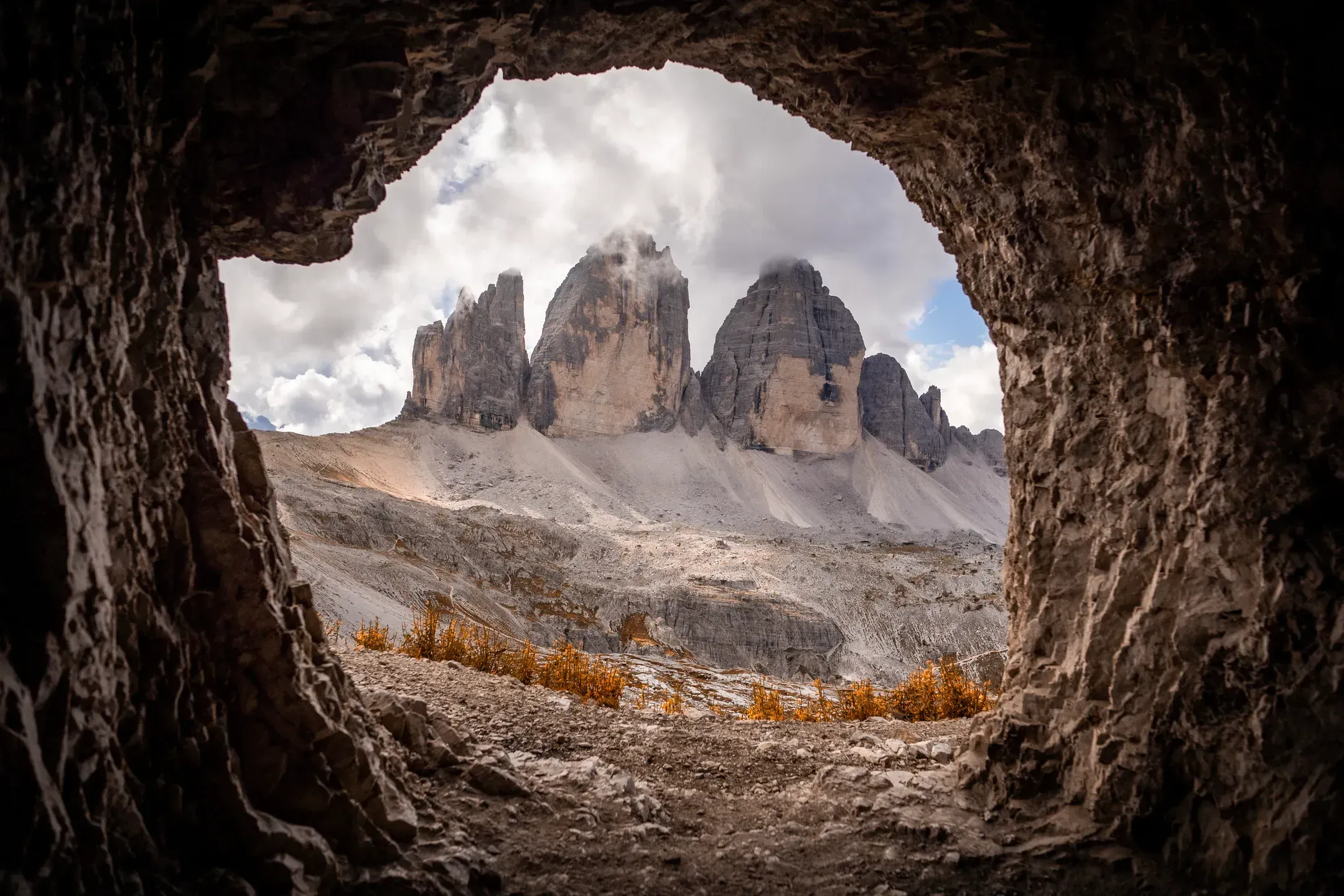 Blick aus einer Felshöhle auf die Drei Zinnen unter Wolken, davor ein Geröllhang mit orangefarbenem Gras