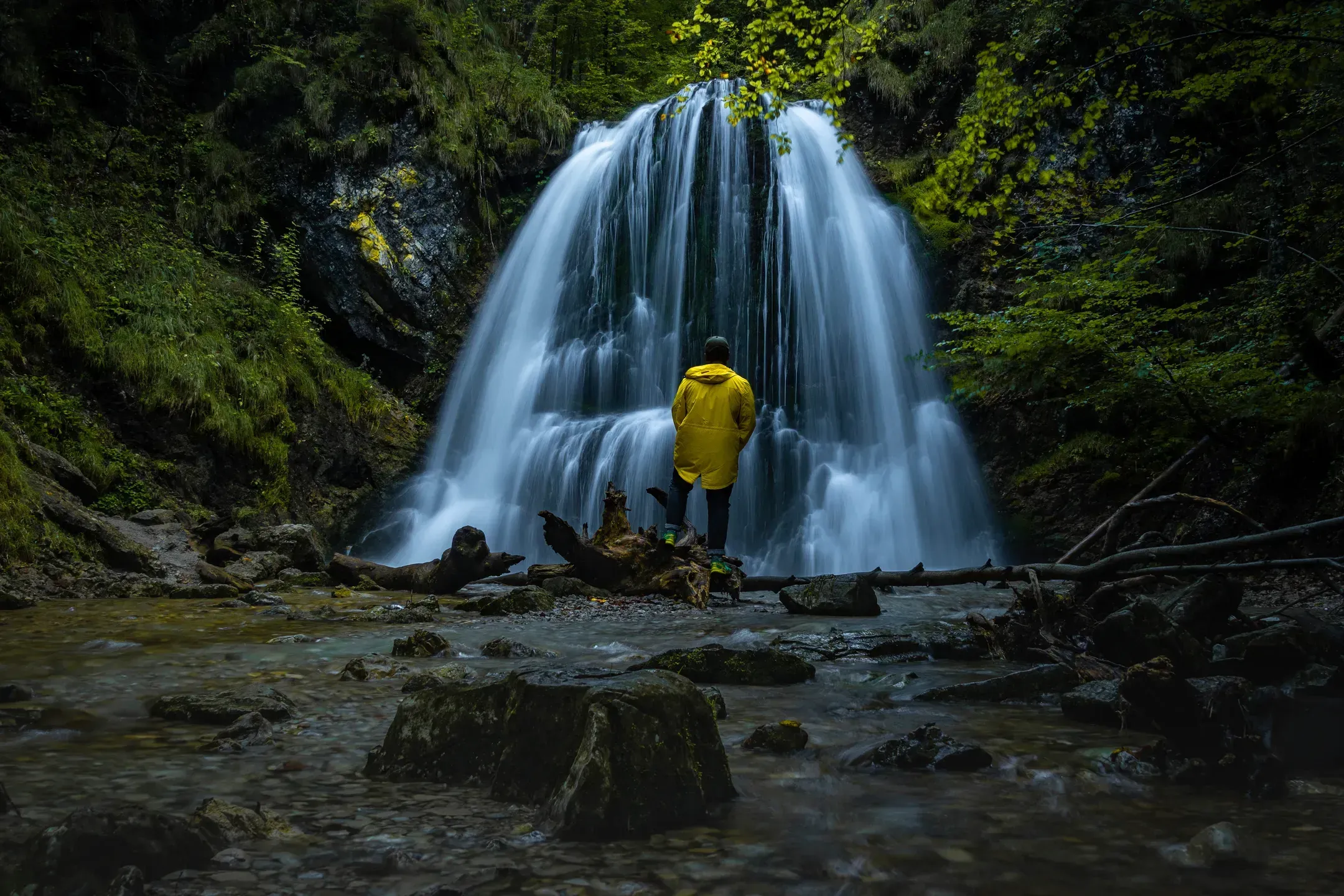 Person in gelber Regenjacke steht vor einem breiten Wasserfall in einer grünen Schlucht, davor ein steiniges Bachbett