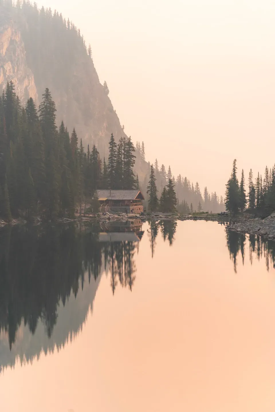 Holzhütte am Ufer eines Bergsees spiegelt sich im glatten Wasser, rosiger Dunst über Wald und Felswand, Hochformat