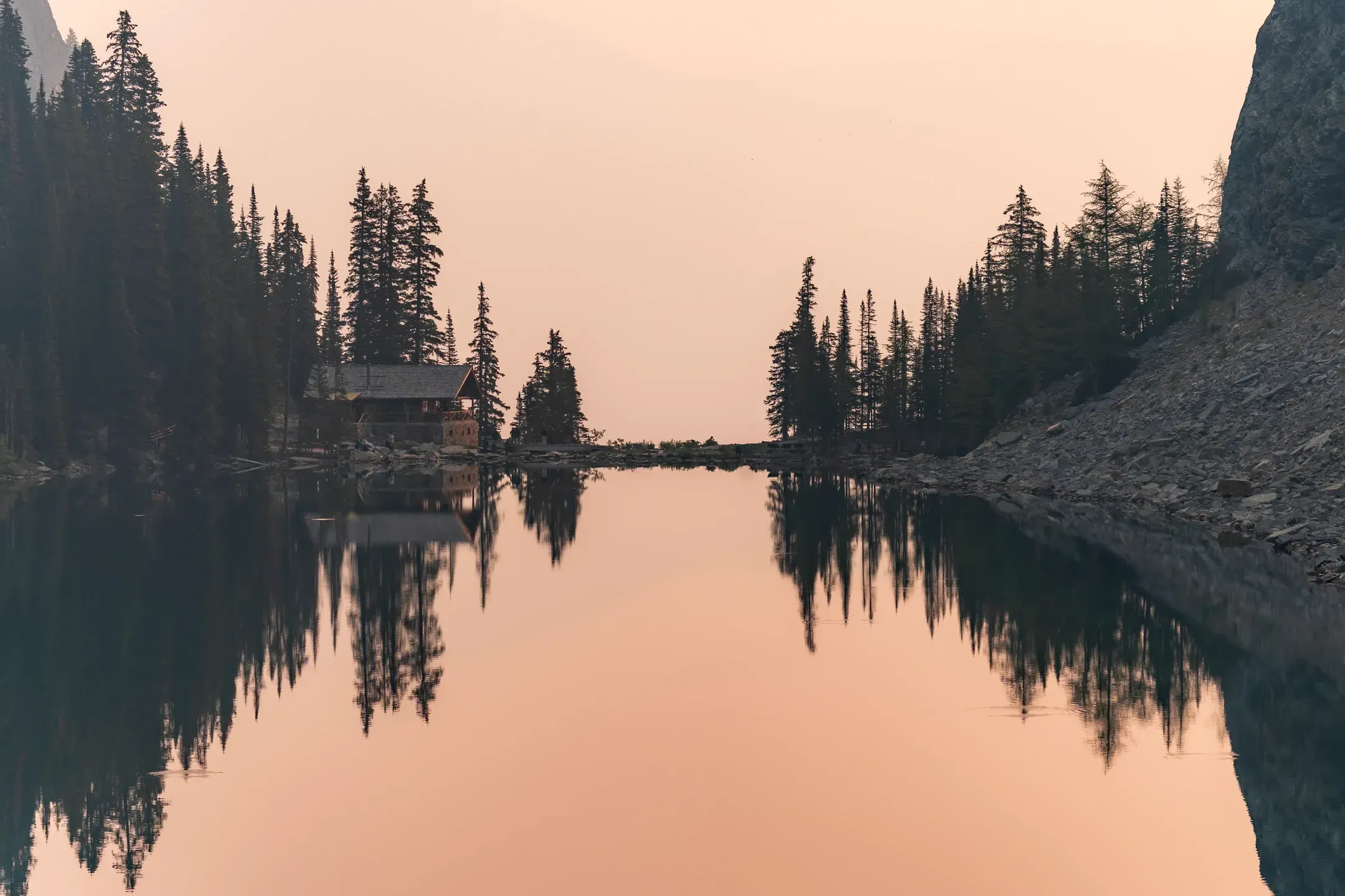 Holzhütte und Nadelbäume spiegeln sich als dunkle Silhouetten in einem stillen Bergsee unter rosa Dunst, Querformat