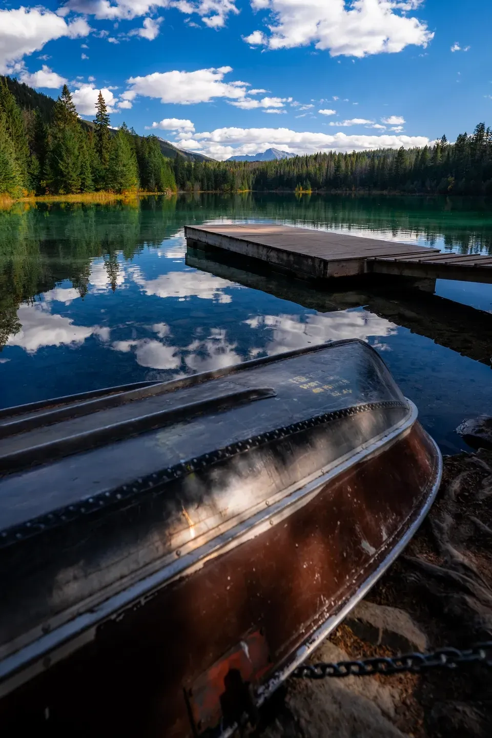 Umgedrehtes Aluminium-Ruderboot am Seeufer, dahinter ein Holzsteg und Wolken, die sich im Wasser spiegeln