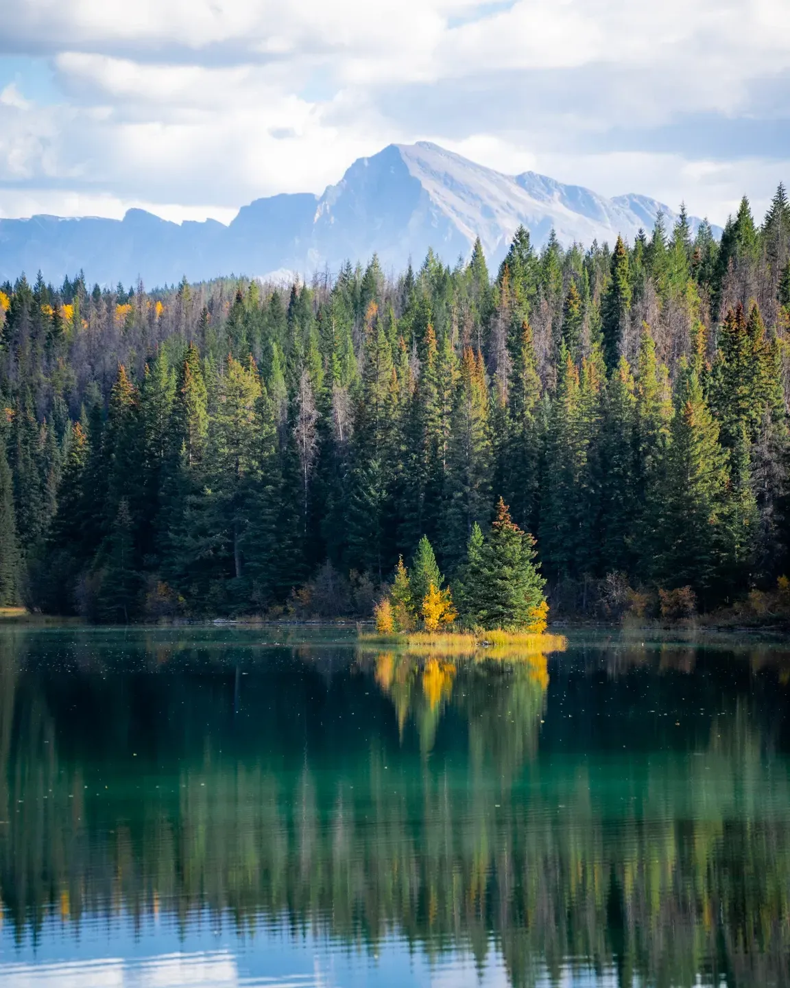 Kleine Insel mit Nadelbäumen und gelbem Herbstlaub spiegelt sich in einem grünen See vor einem hellen Berggipfel
